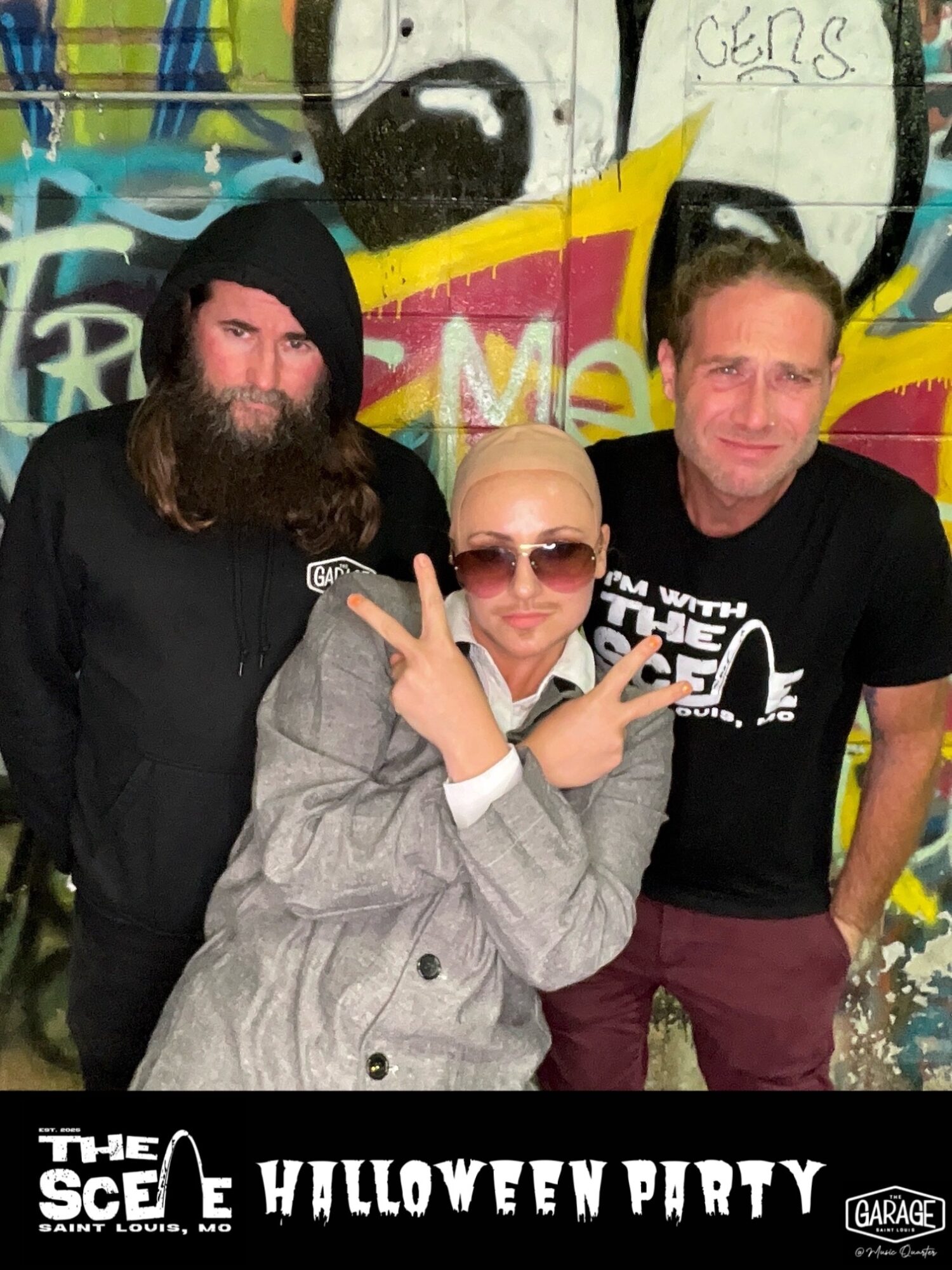 Three people posing in front of a colorful graffiti wall, one making a peace sign, at a Halloween party.