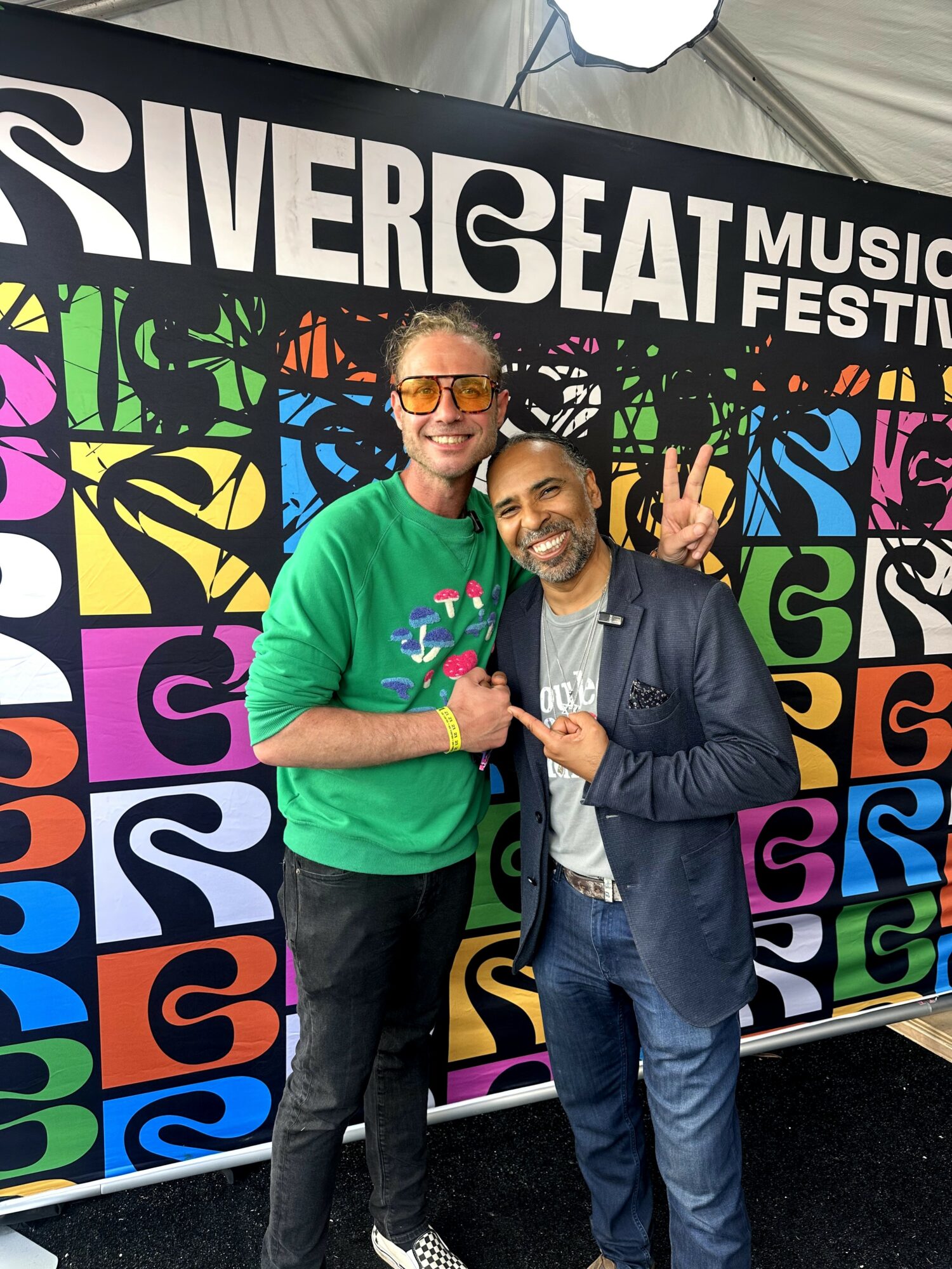 Two people smiling and posing in front of a colorful festival backdrop, one making a peace sign gesture.