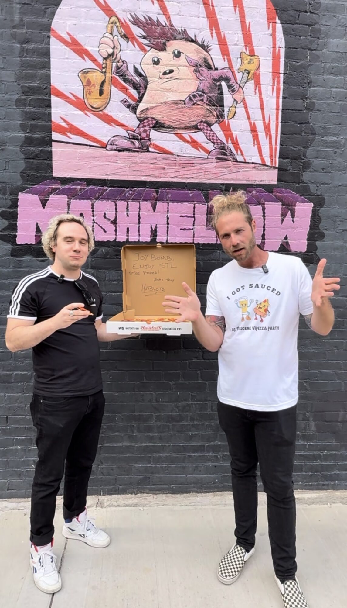 Two men stand in front of a colorful mural with a pig character and the word 'MASHMEOW'. One holds a pizza box.