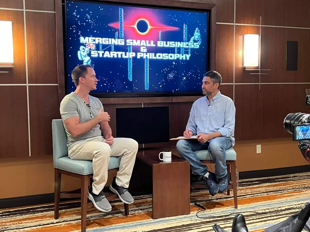 Two people sit and talk in a room with a large screen displaying a presentation about merging small business and startup philosophy.