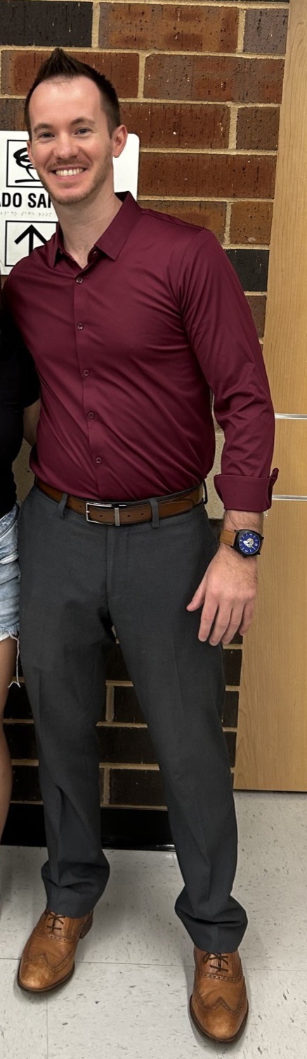 Man with short hair and beard wearing a maroon shirt, dark pants, and brown shoes, standing indoors against a brick wall.
