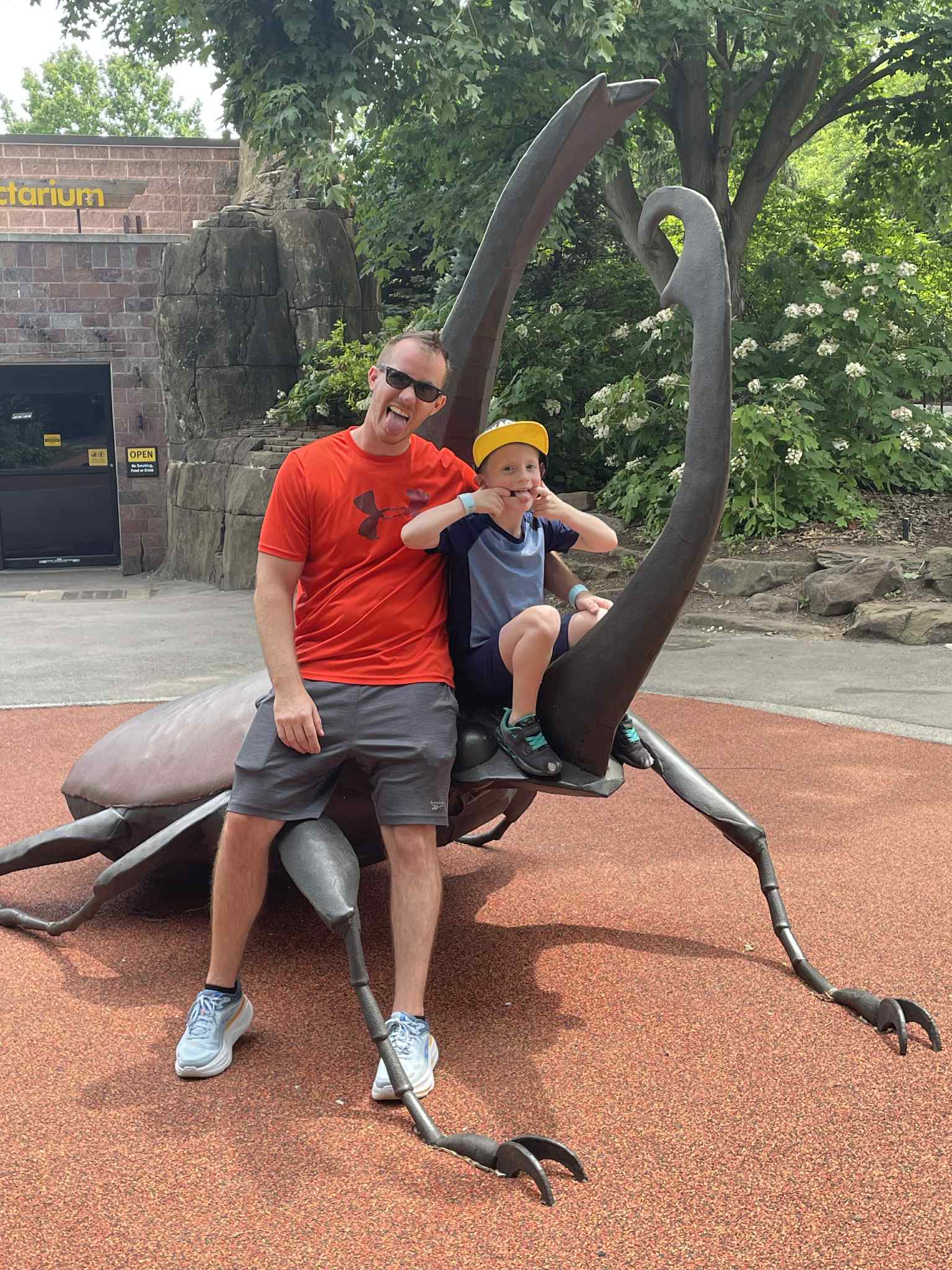 Man and child sitting on a large insect sculpture outdoors, surrounded by trees and greenery.