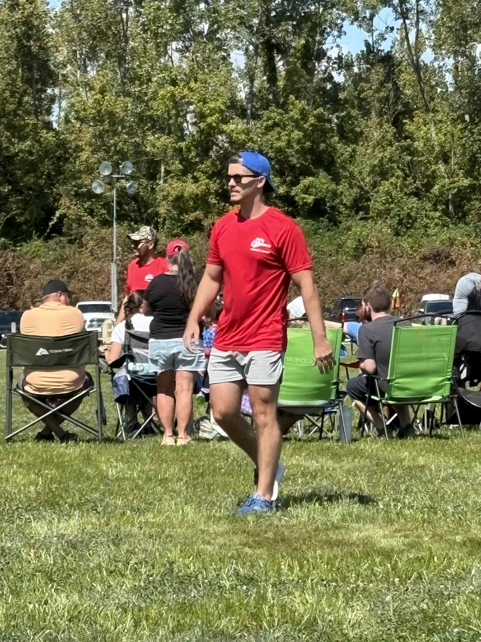 Man in red shirt, gray shorts, blue cap, sunglasses walking on grass during outdoor event with seated crowd and trees.