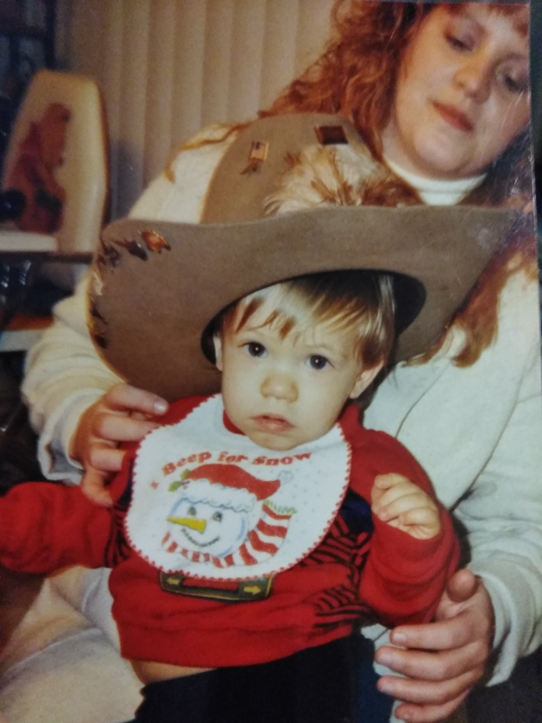 Child wearing a large hat and a festive bib, held by an adult woman with curly hair, indoors.