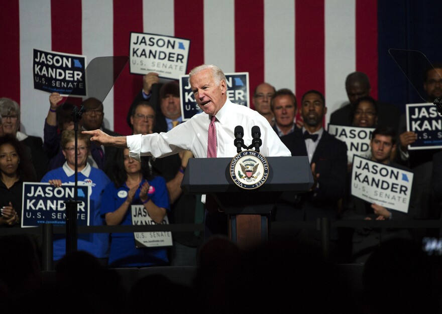 Joe Biden speaking at a podium with a group of people holding campaign signs behind him.