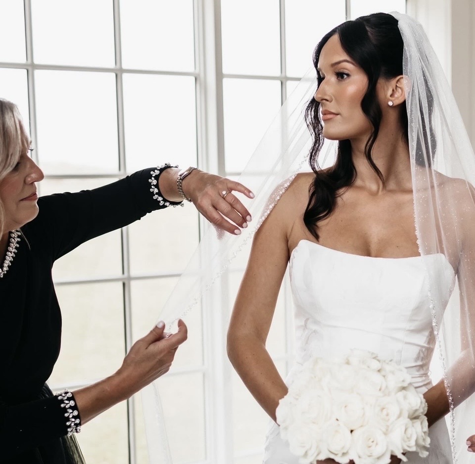 Bride in white dress with veil, standing indoors, facing right, with woman adjusting her dress or veil.