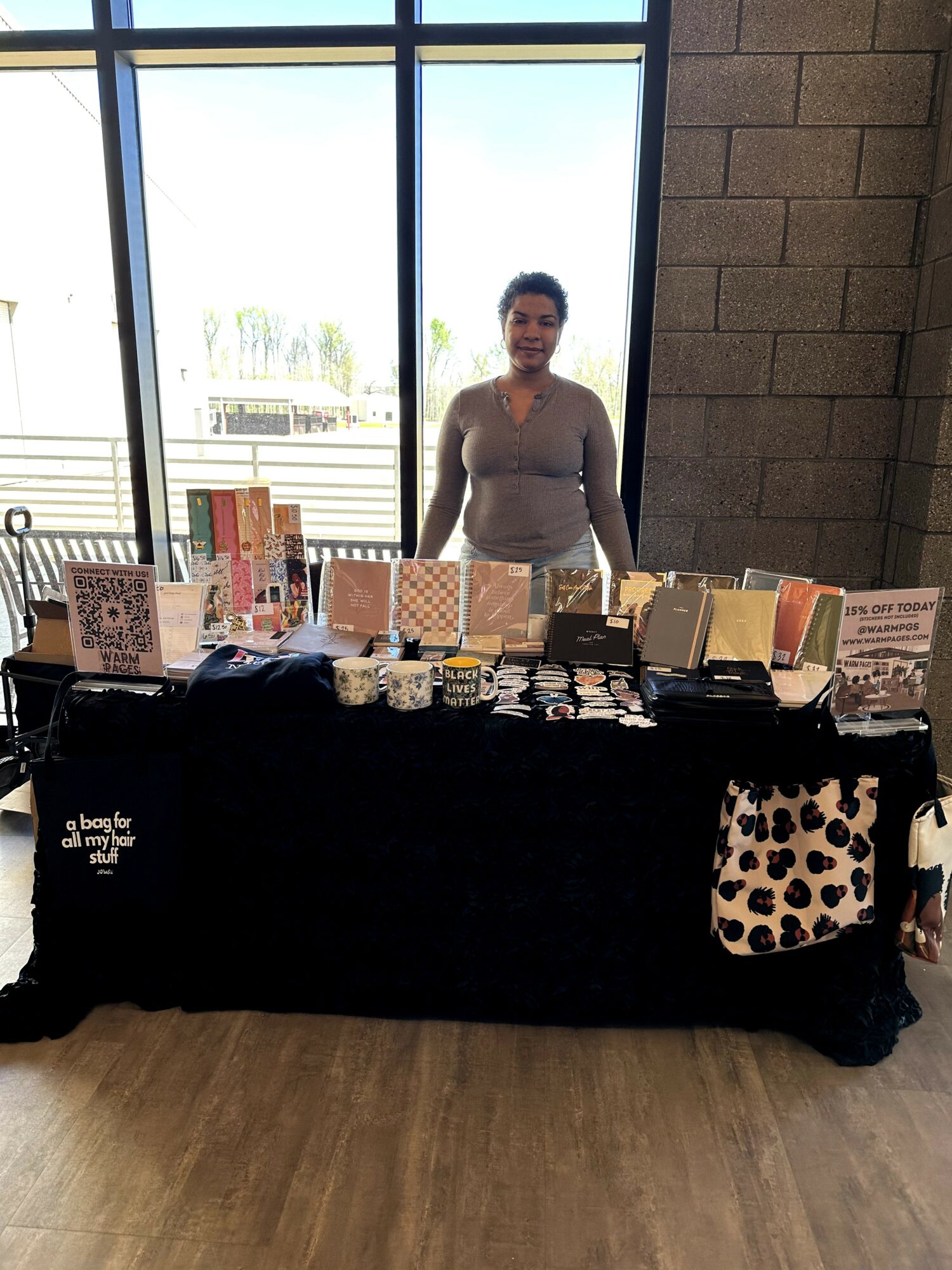 Woman standing behind a table with books, cards, and items inside a building with large windows.