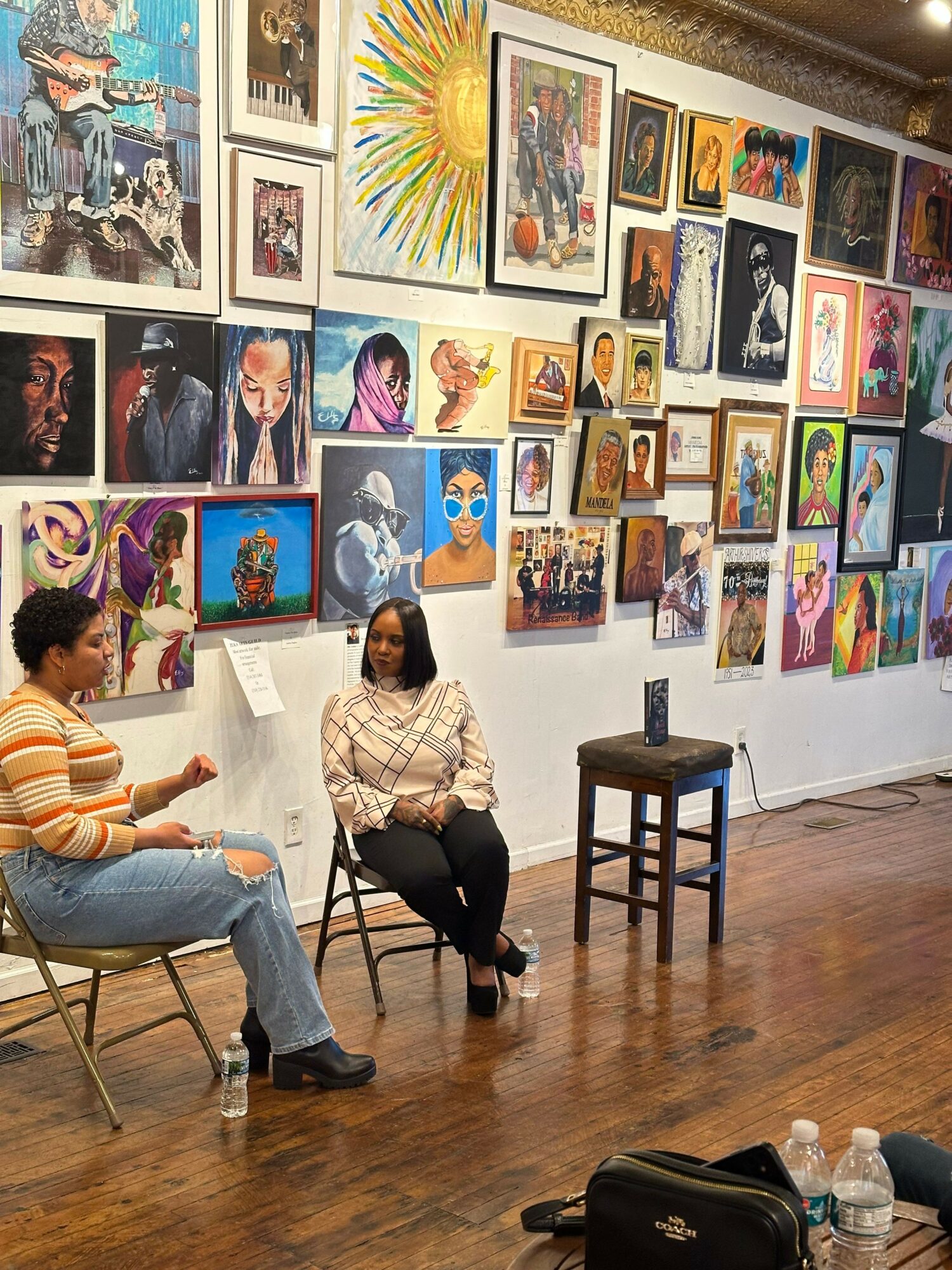 Two women sit and talk in an art gallery with colorful paintings on the wall behind them.