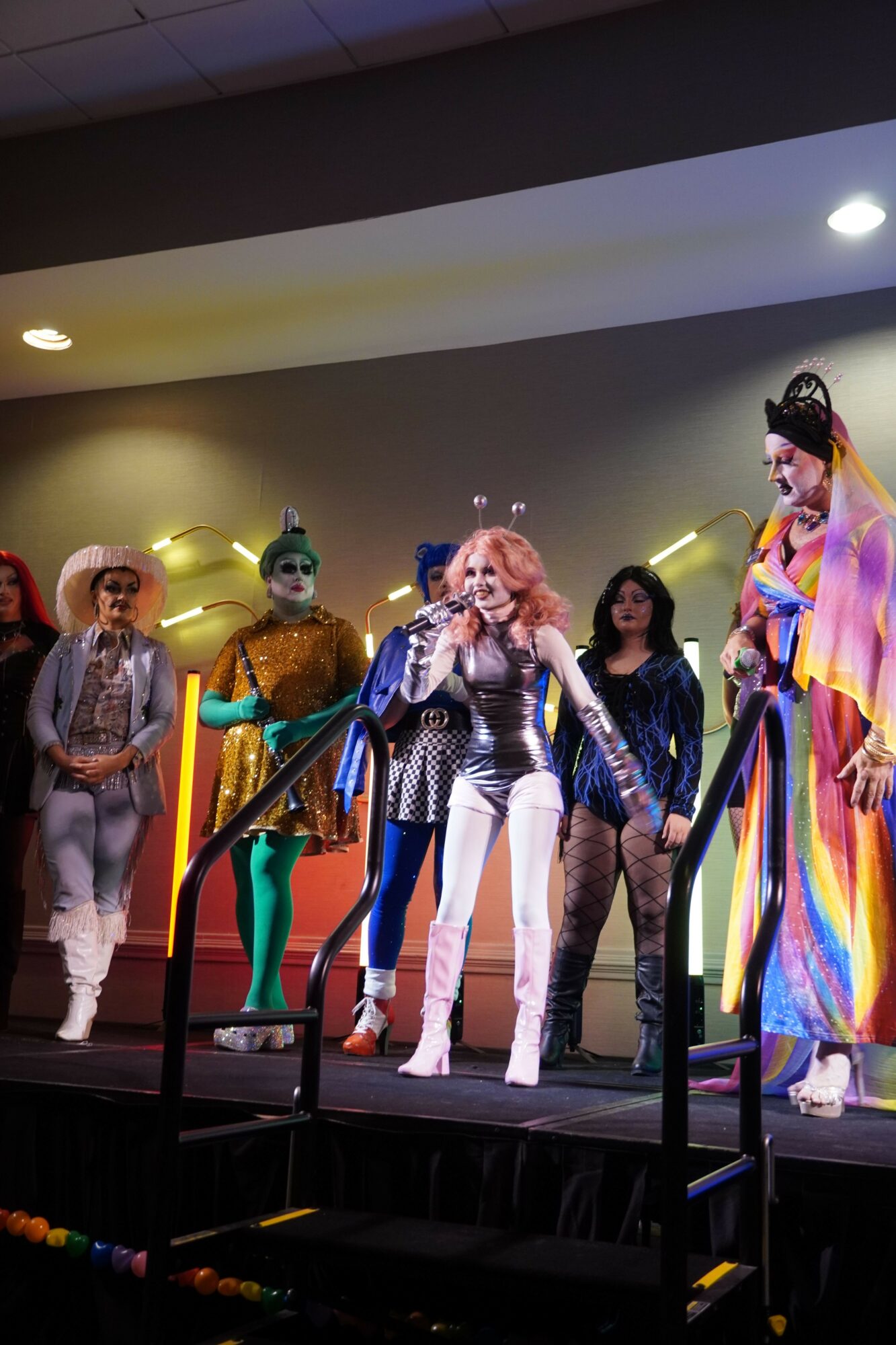Group of people in colorful costumes on stage, some with masks and wigs, performing or posing.