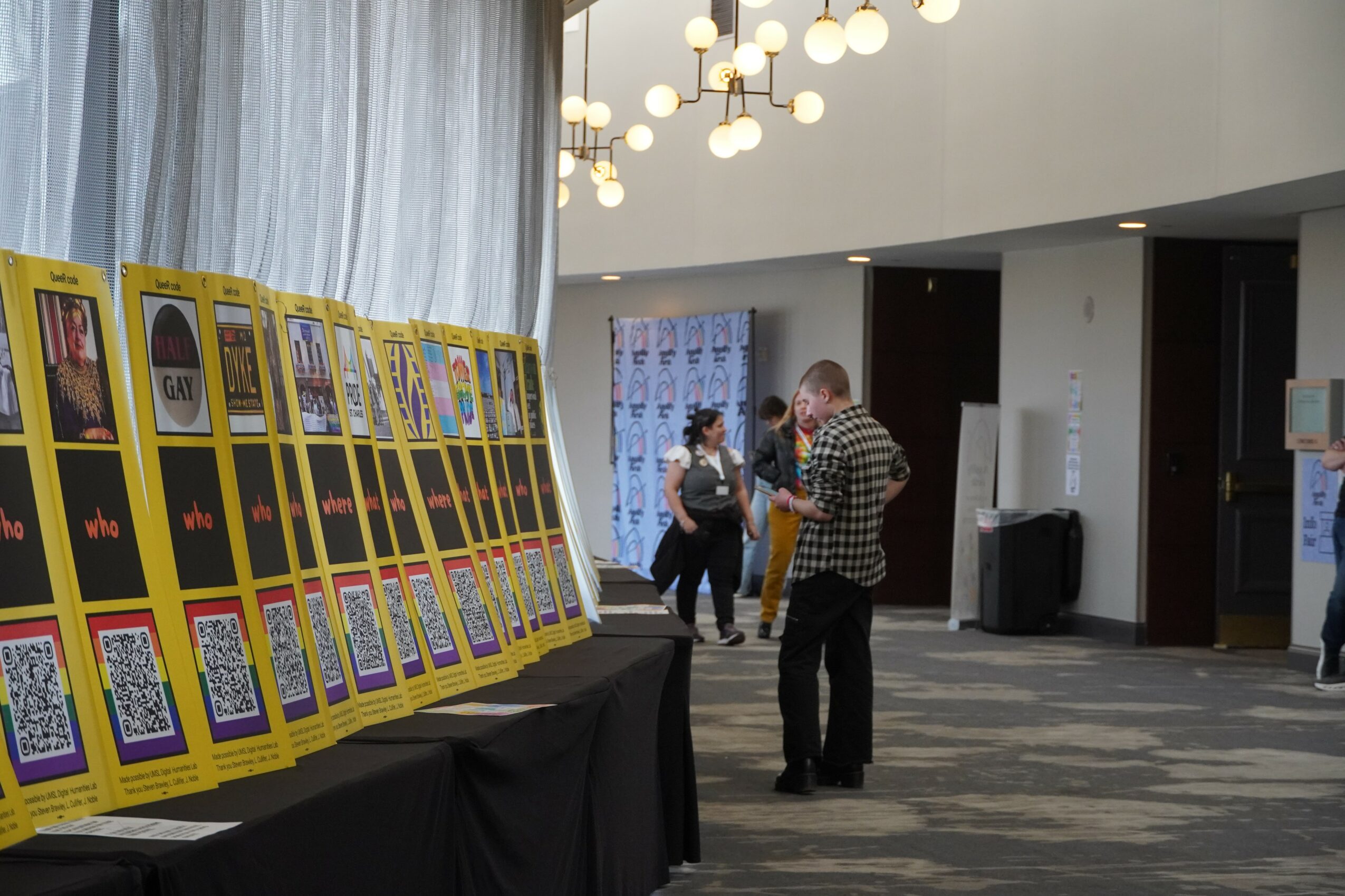 People in a lobby area looking at large yellow display boards with QR codes and images, with a table nearby and modern lighting.