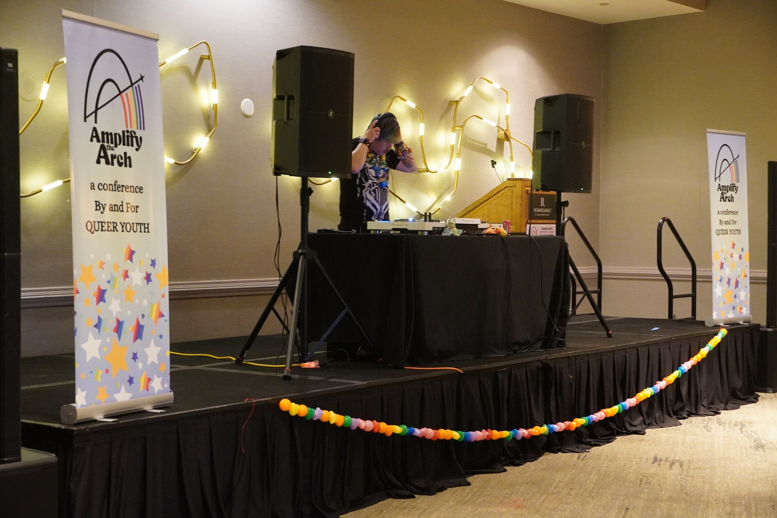 DJ performing on stage with speakers and banners at an indoor event, decorated with string lights and colorful garland.