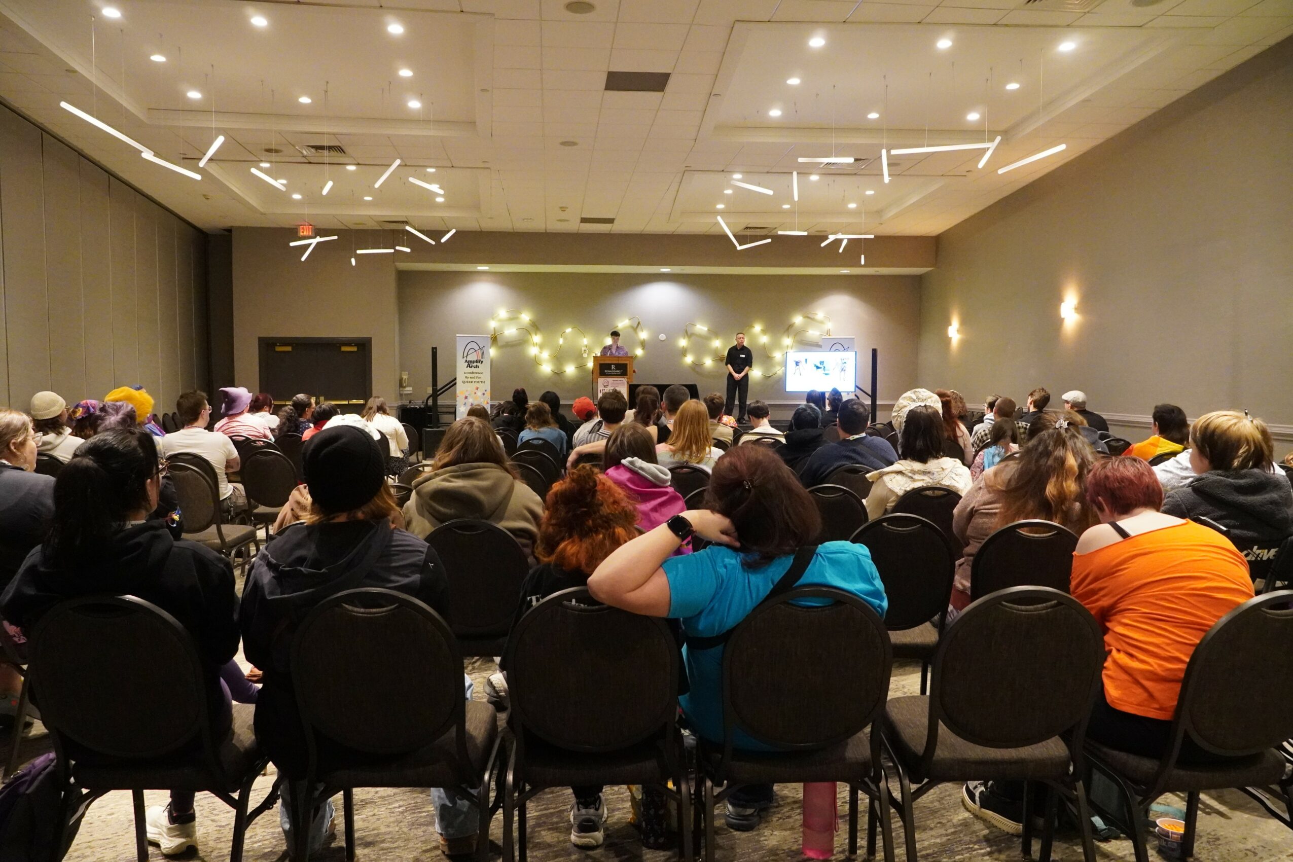 Large audience seated in a conference room facing a stage with speakers and a presentation screen.