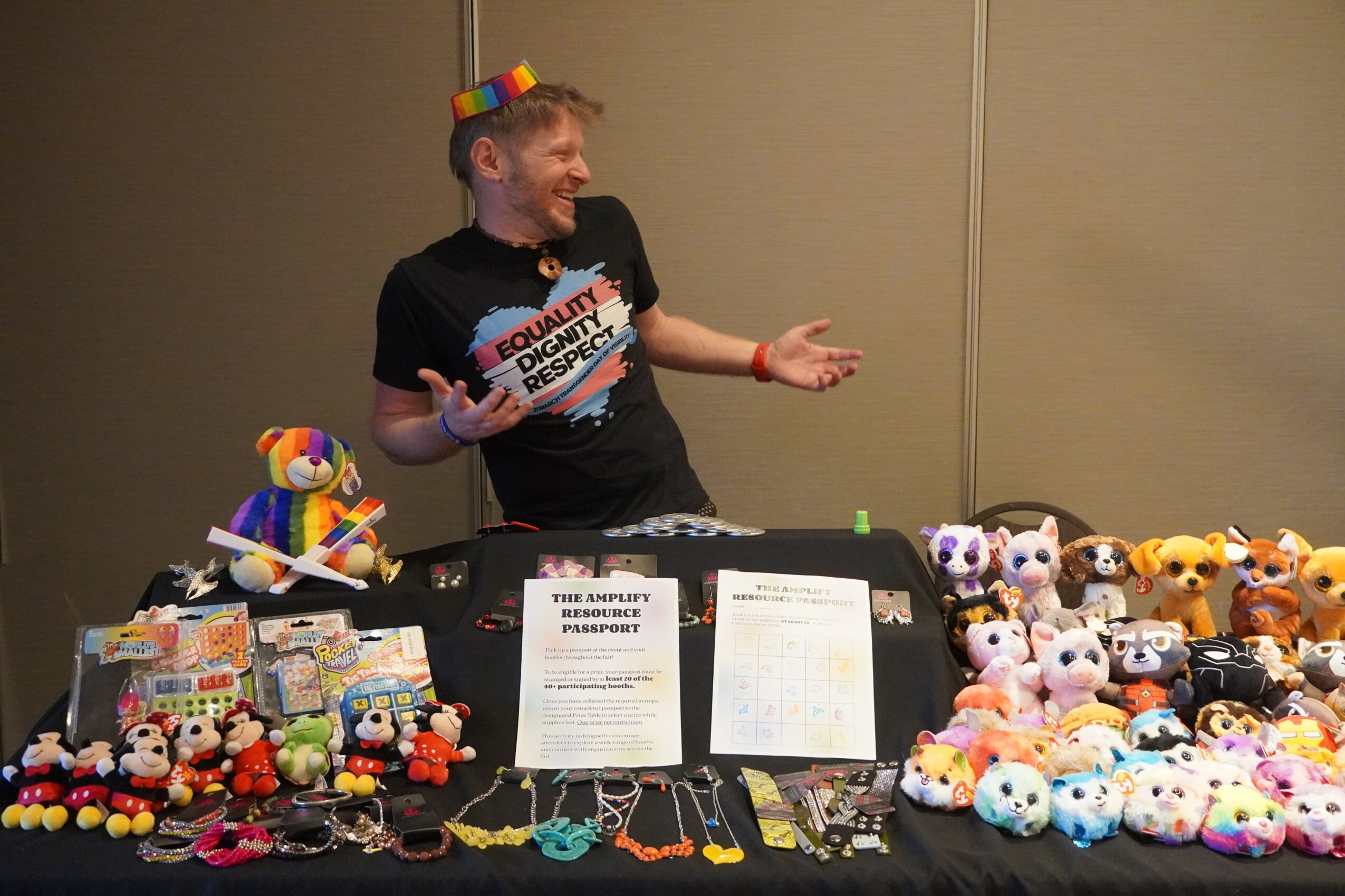Person wearing a black t-shirt with colorful text, smiling and gesturing, surrounded by plush toys and items on a table.