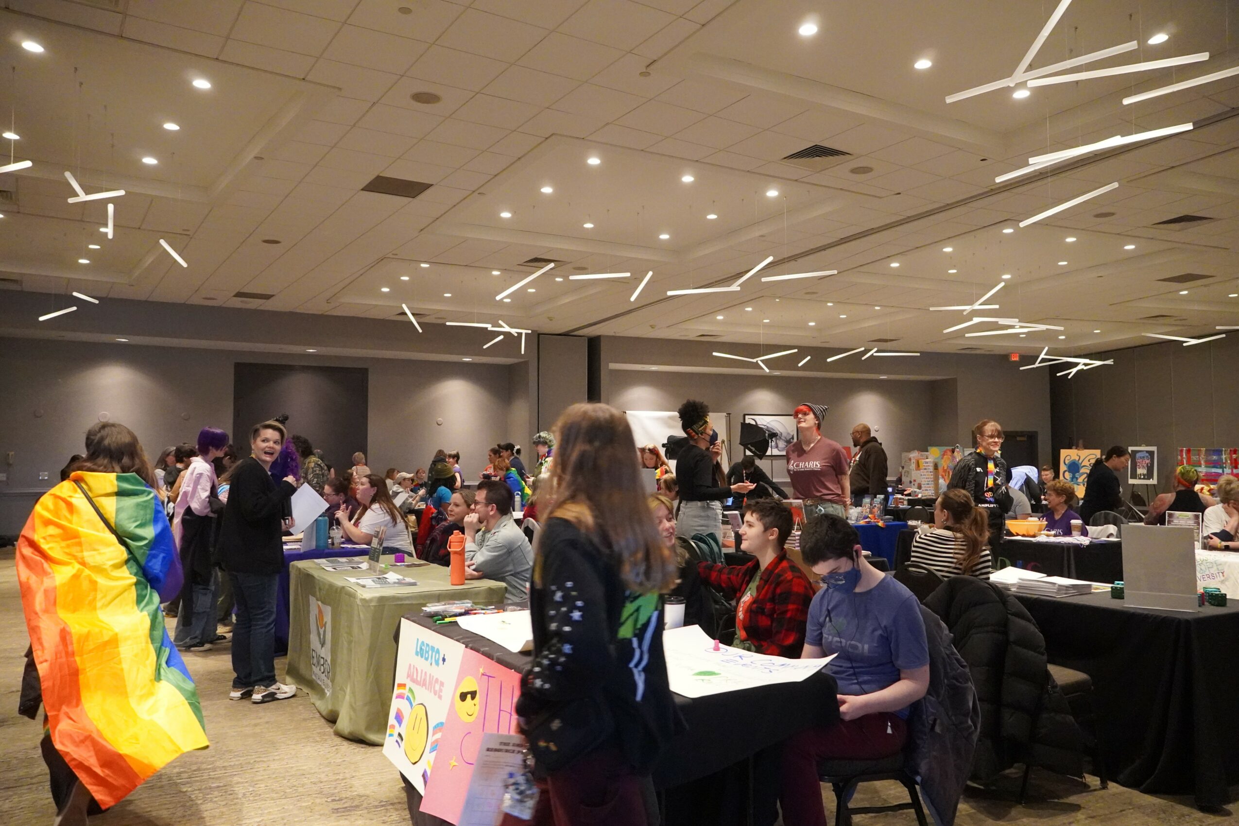 People gather in a large indoor space with tables and displays, some wearing colorful clothing, engaging in activities.