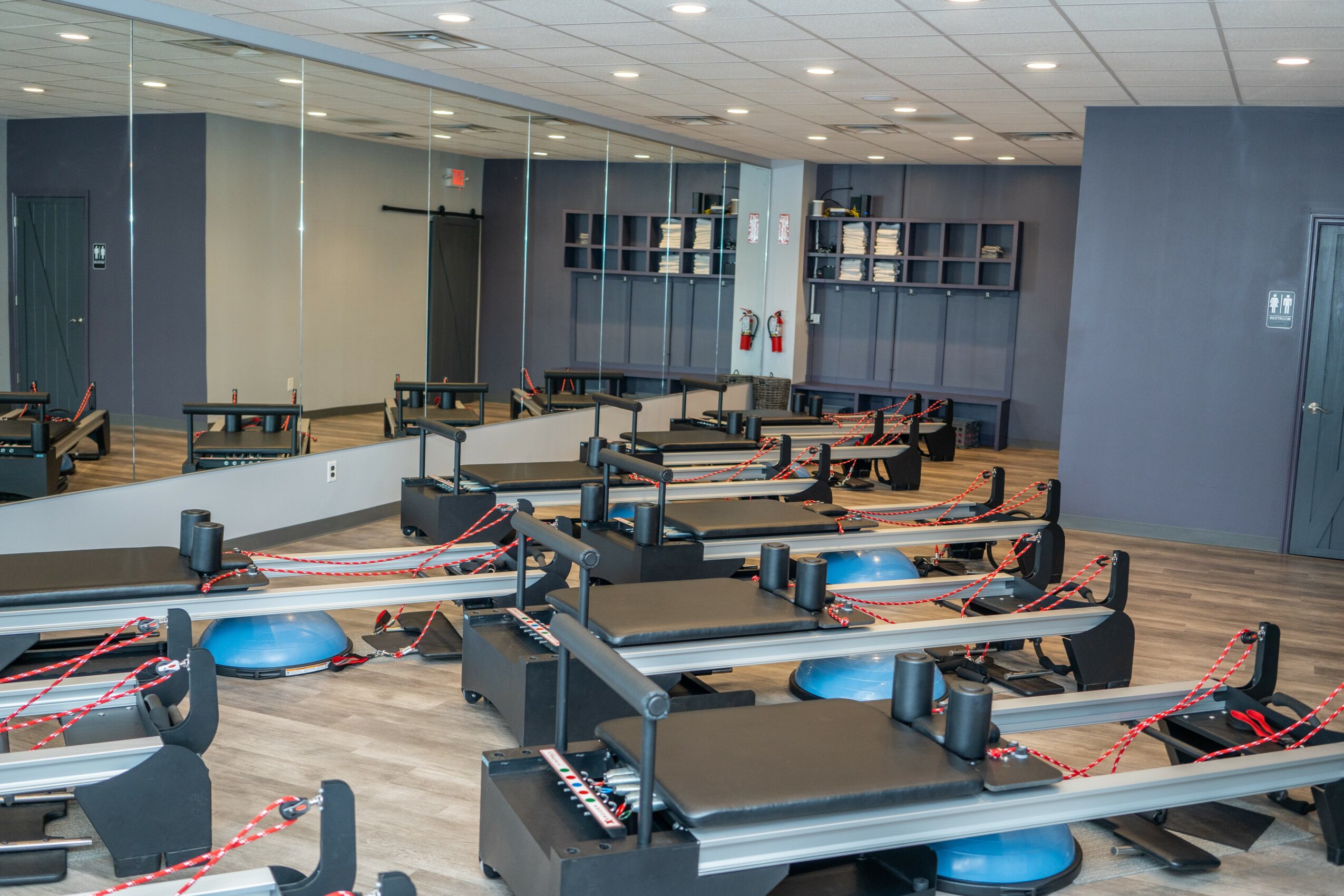 Fitness room with exercise benches, mats, and equipment, separated by red and white striped tape, with mirrors and storage shelves.