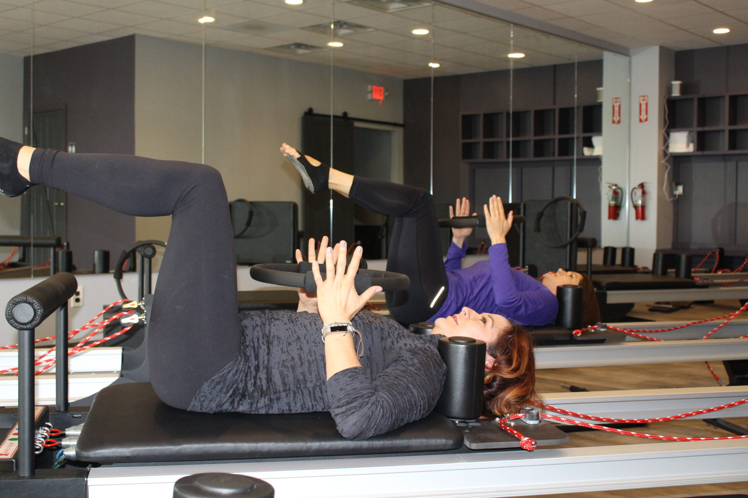 Person lying on a mat with legs raised and hands near head in a fitness class, others exercising in background.