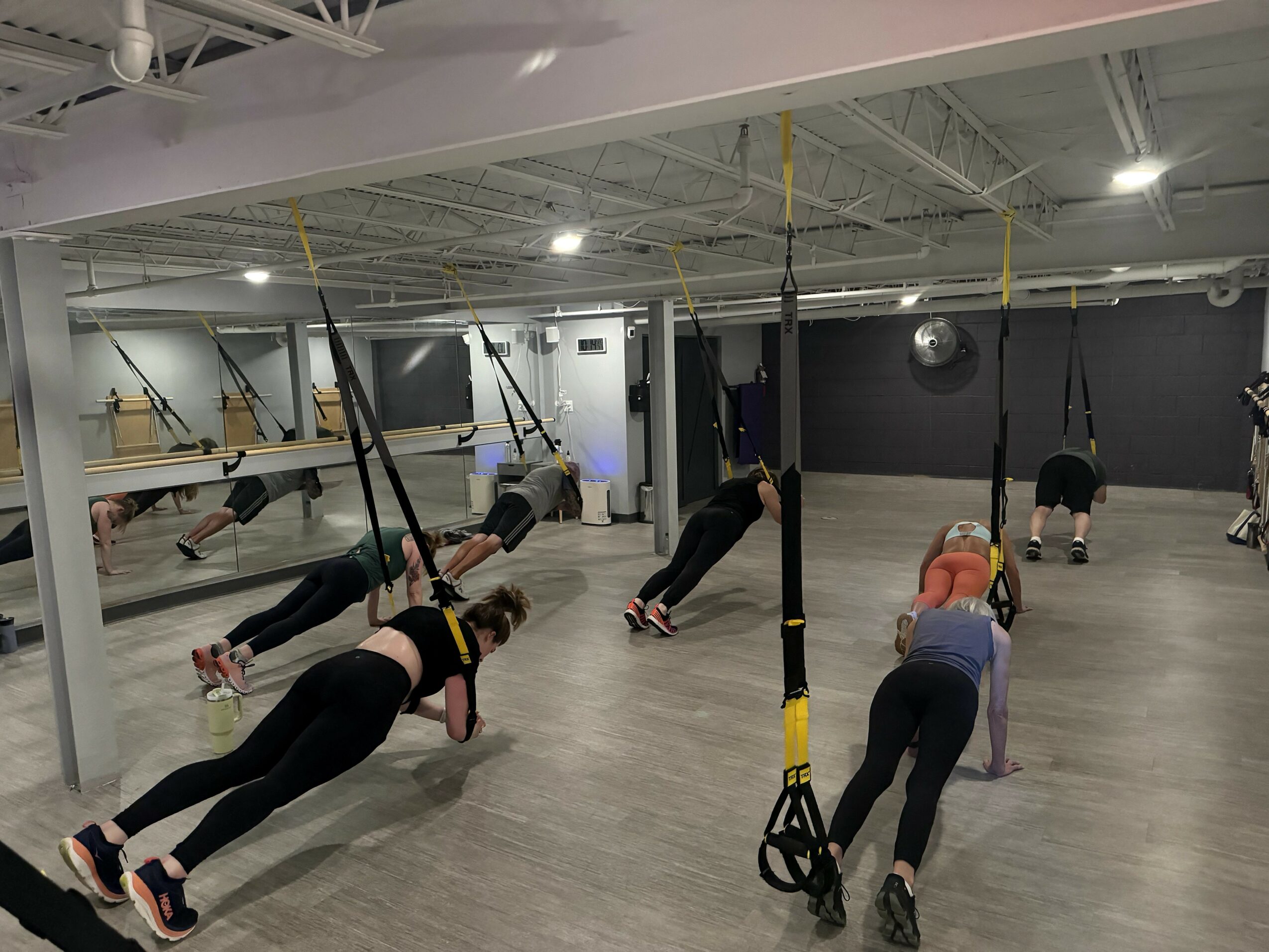 People exercising with suspension trainers in a fitness studio, some in plank positions, mirrors on wall, wooden floor.