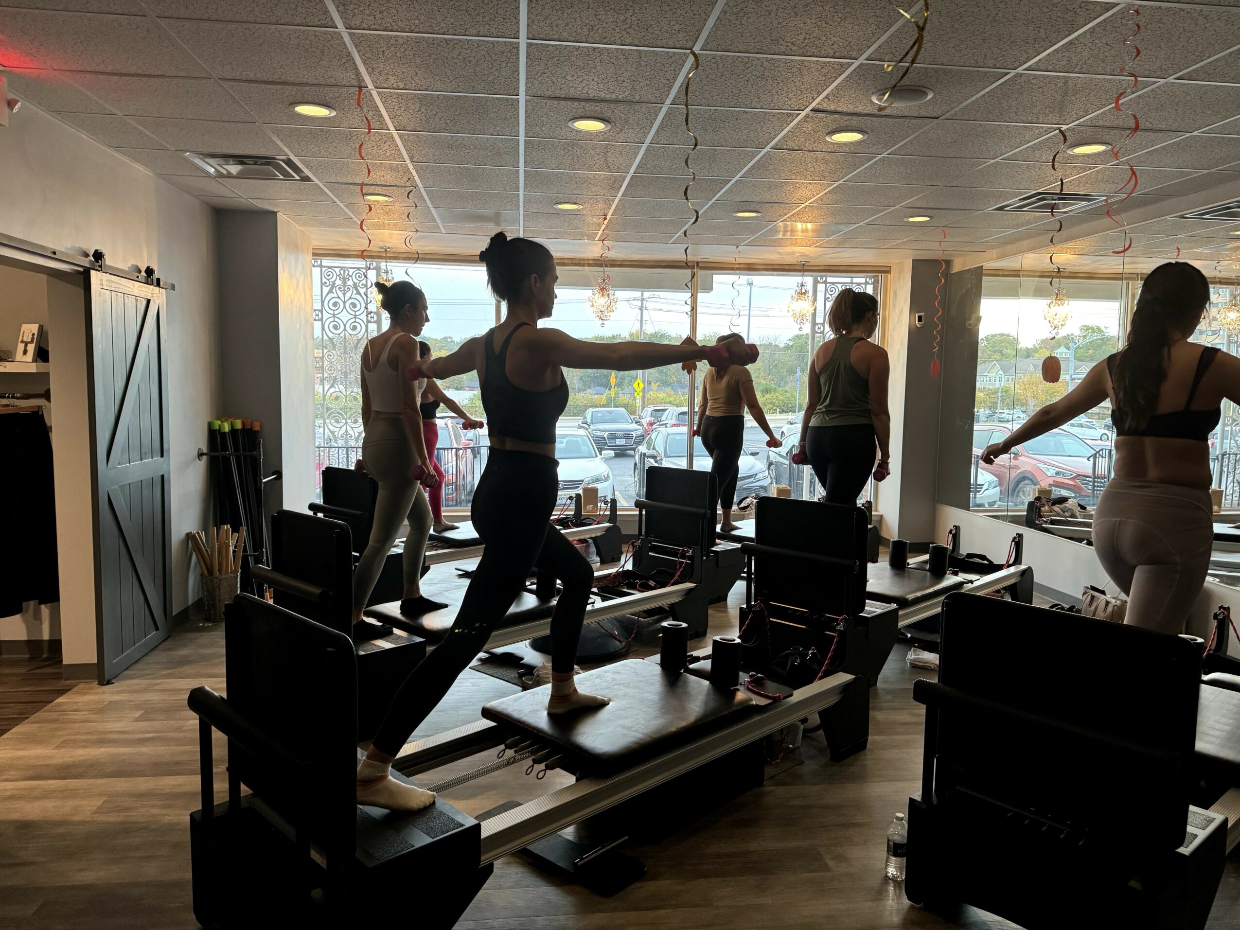 People practicing Pilates on reformer machines in a fitness studio with large windows and wooden floors.