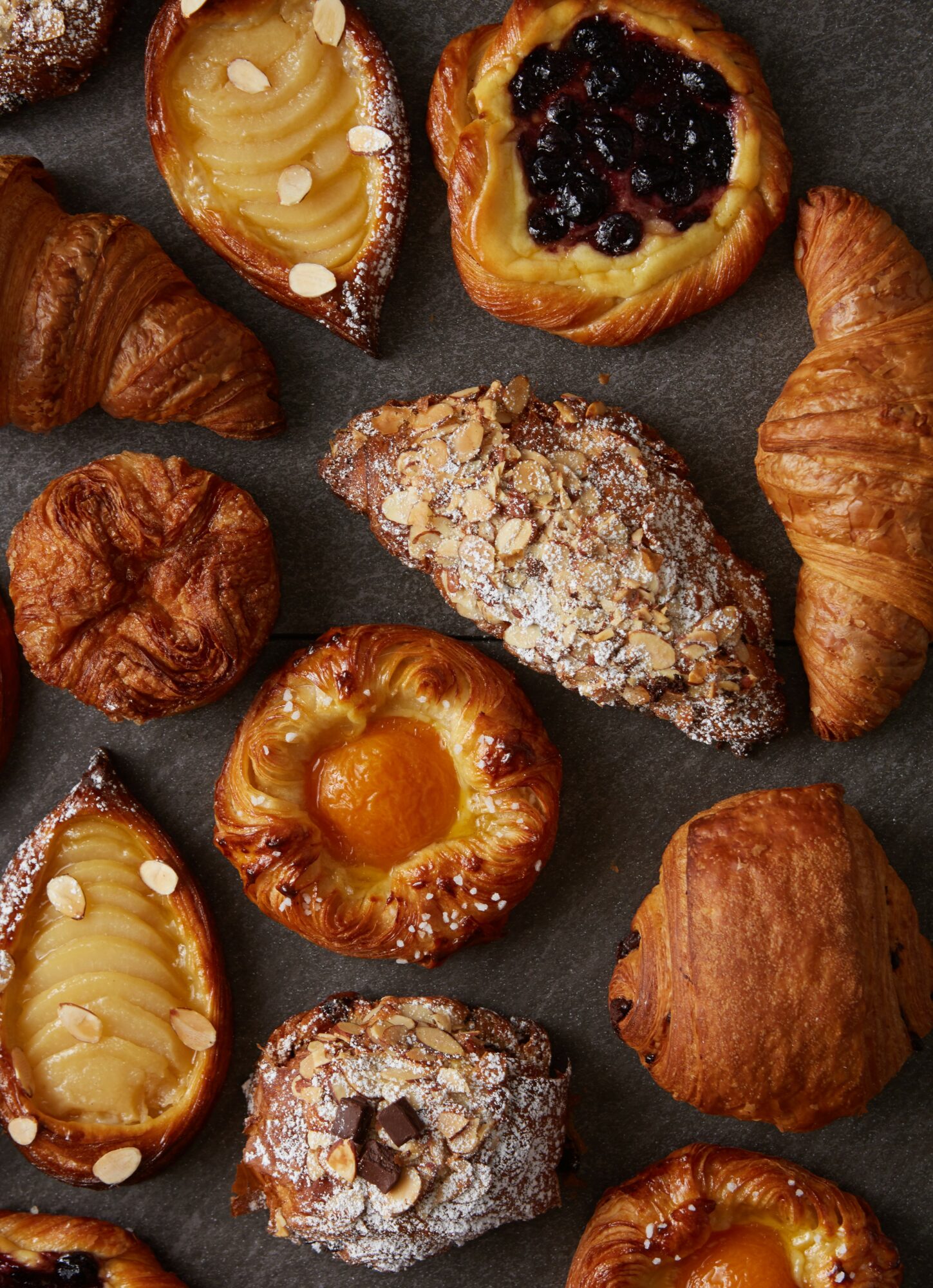 Assorted pastries including croissants, Danish pastries with fruit filling, and powdered sugar-covered treats on a dark surface.