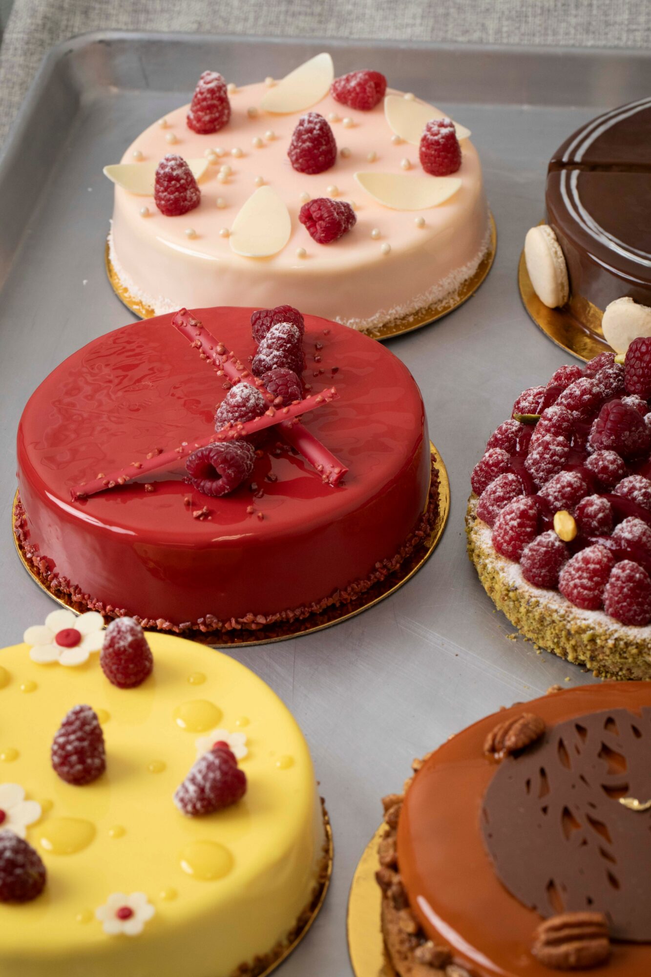 Four decorated cakes with berries and chocolate on a tray, including pink, red, yellow, and chocolate flavors.