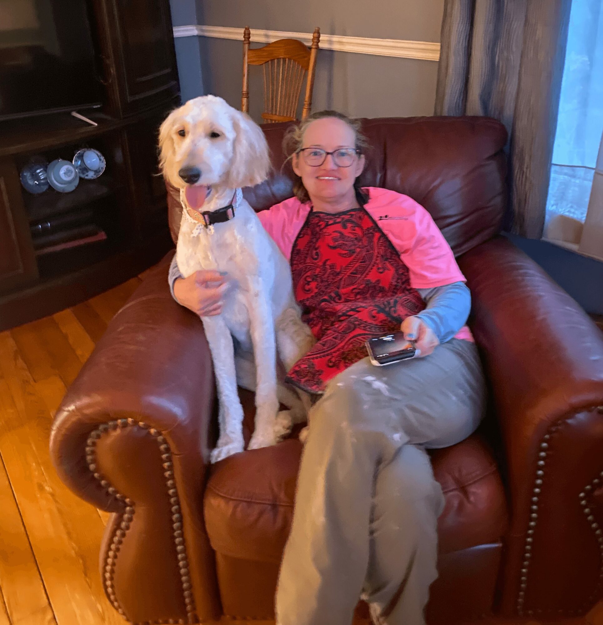 Person with glasses sitting on a brown leather couch, holding a large white dog, in a living room.