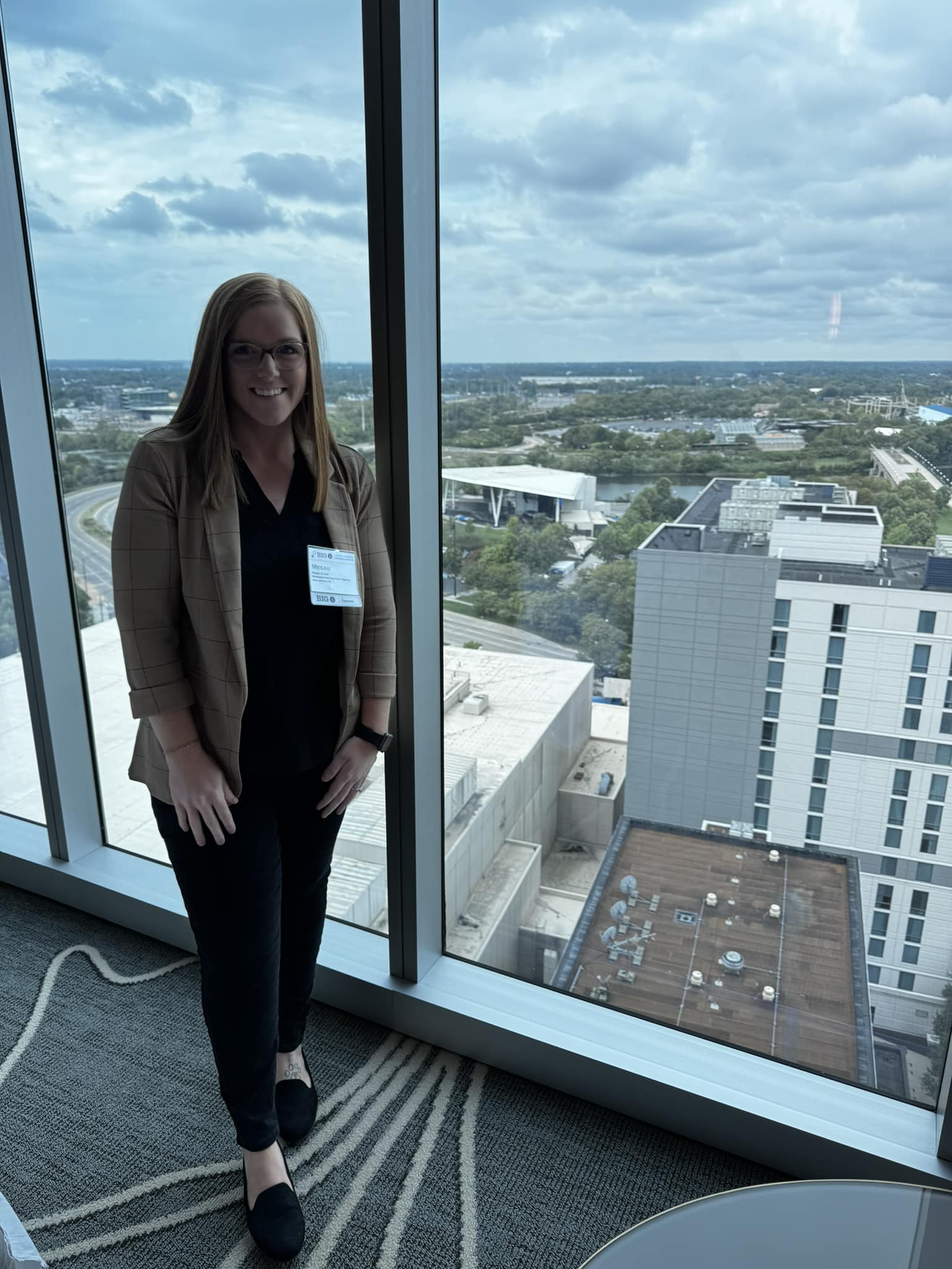 Woman standing indoors near large window overlooking cityscape and sky.