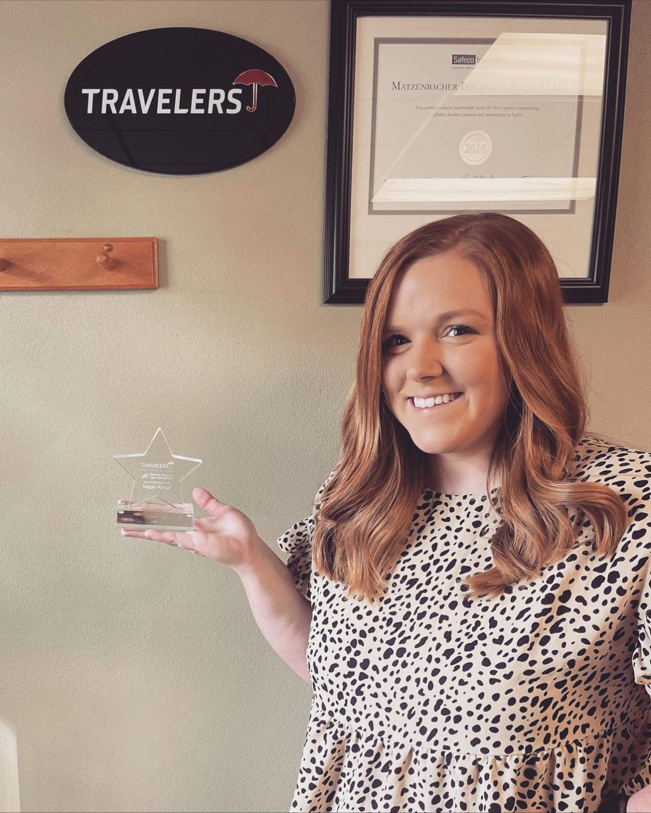 Woman with long hair holding a star-shaped award, smiling, in front of framed certificates and a 'TRAVELERS' sign.