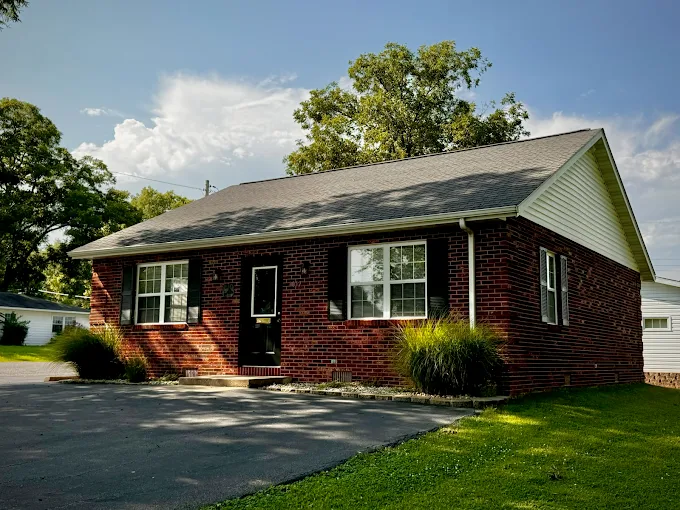 Brick house with white trim, black door, and large windows, surrounded by green grass and trees under a partly cloudy sky.