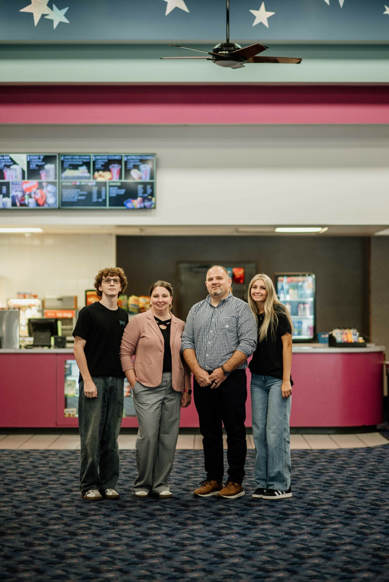 Four people standing in front of a counter in a colorful indoor setting, with screens and decorations above.