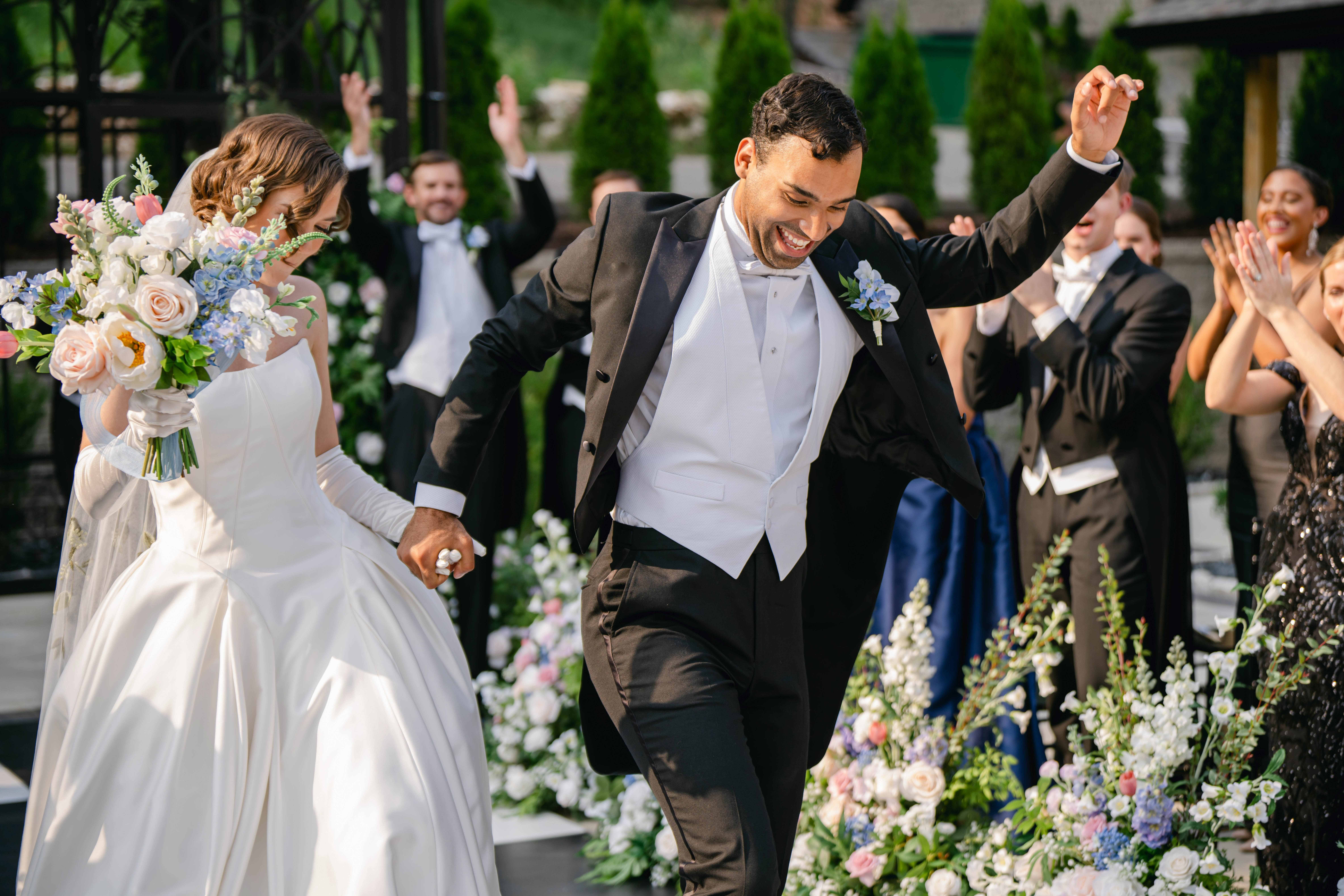 Man in tuxedo celebrating with a woman in wedding dress outdoors, surrounded by people and floral decorations.