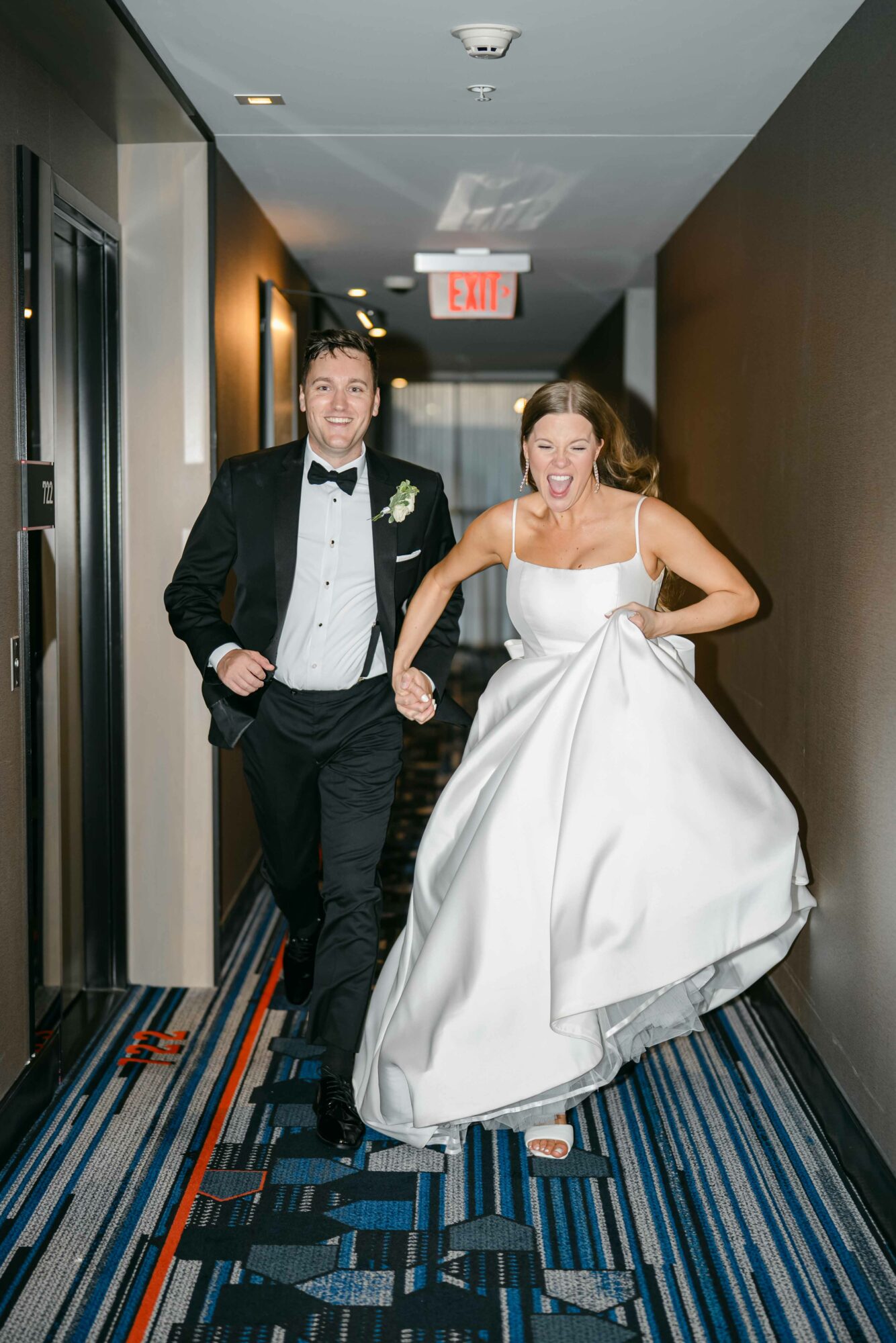 A man in a tuxedo and a woman in a white gown walk down a hallway holding hands.