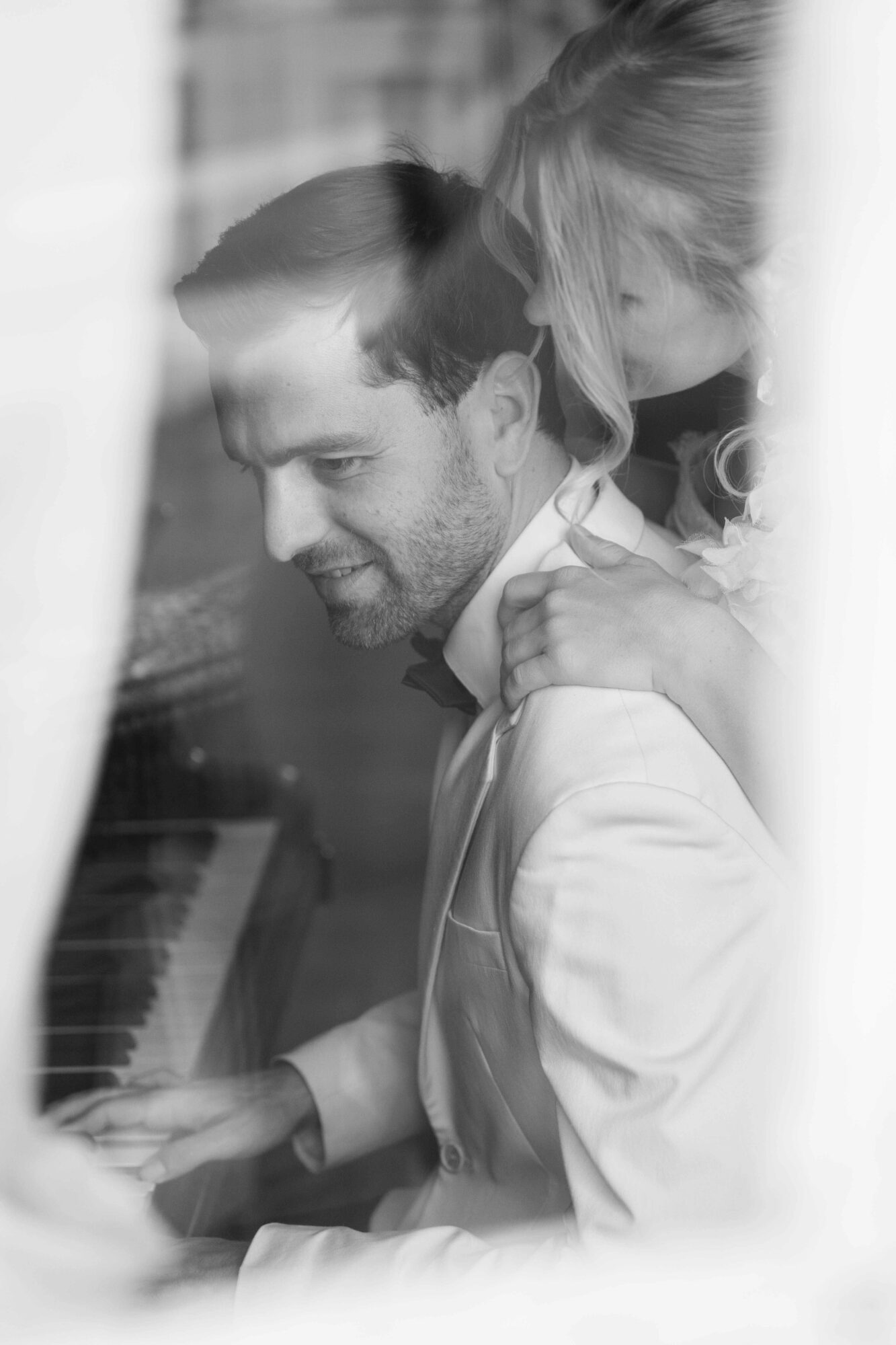 Two people, one playing piano and the other leaning on their shoulder, seen through a window or glass.