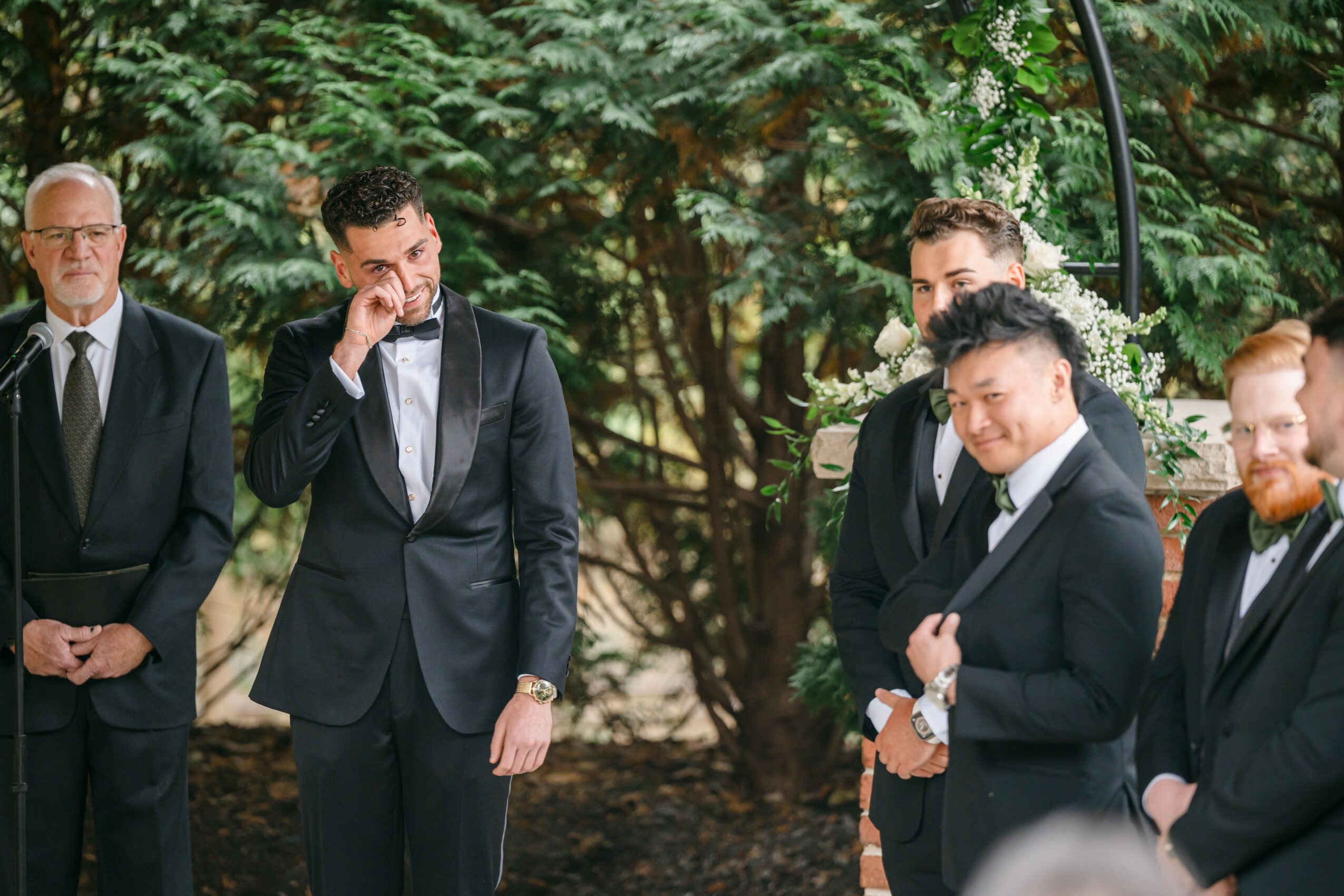 Group of five men in suits standing outdoors among trees, some smiling and talking.