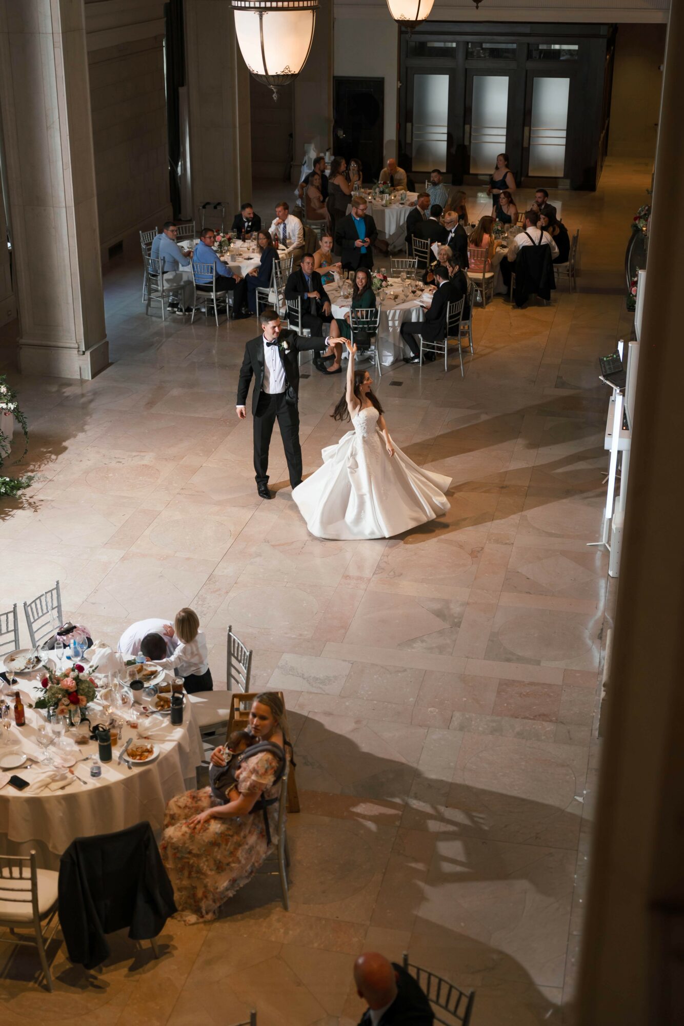 Wedding reception with bride and groom dancing, guests seated at tables, and a large chandelier overhead.