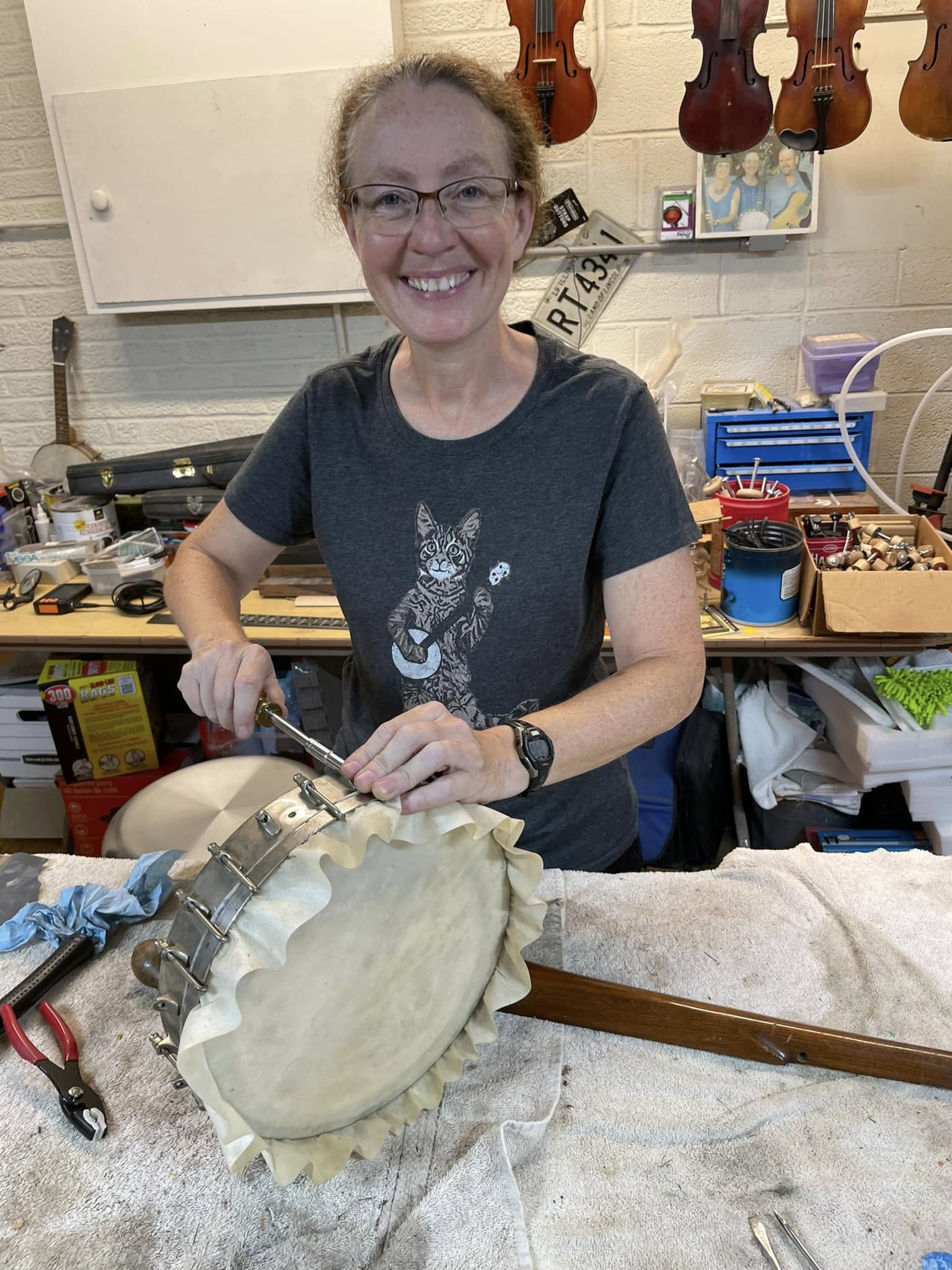 Smiling woman with glasses working on a musical instrument in a workshop, surrounded by tools and musical instruments.