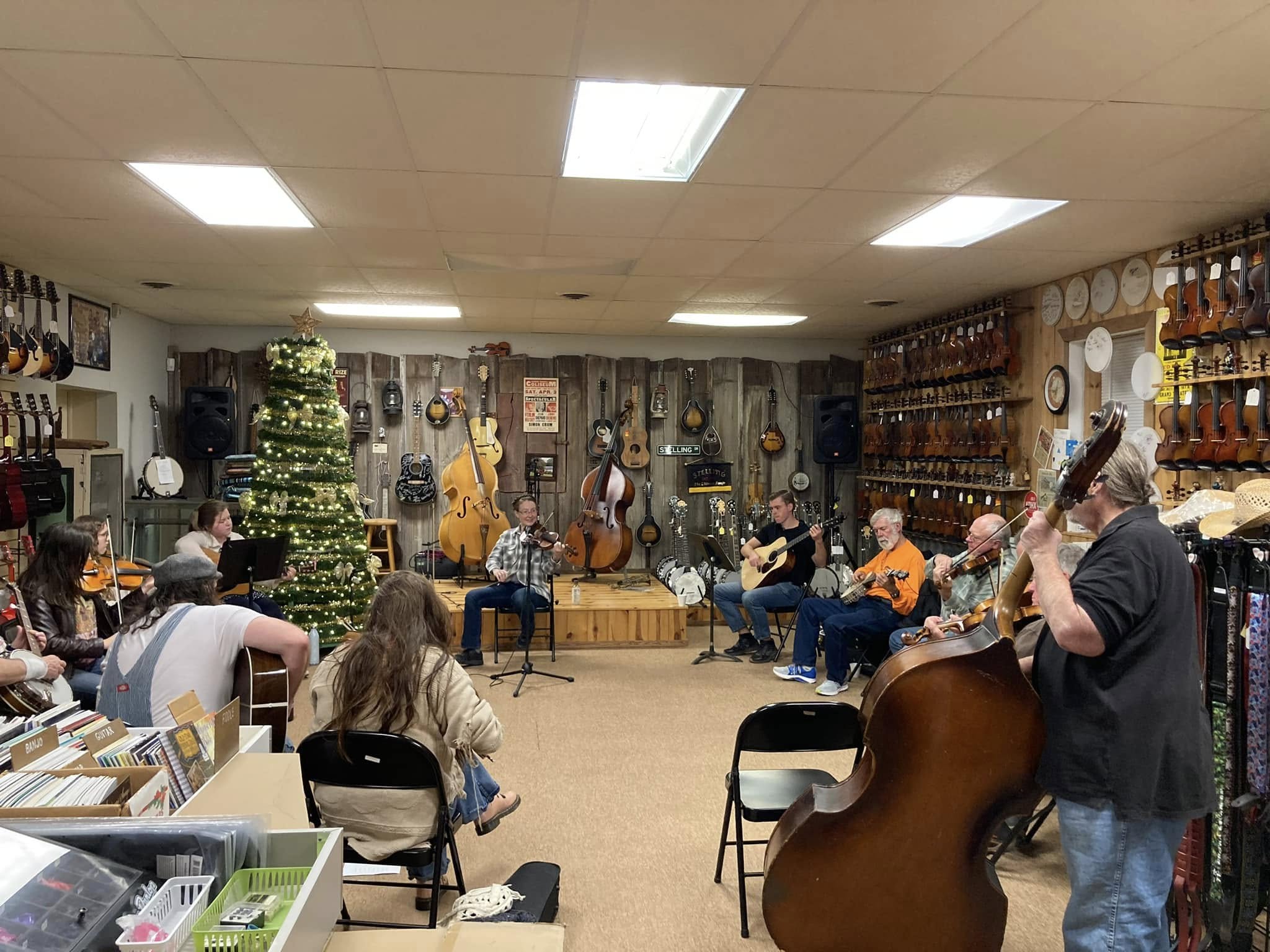 Group of people playing musical instruments in a room decorated with guitars and ornaments, with a Christmas tree in the background.