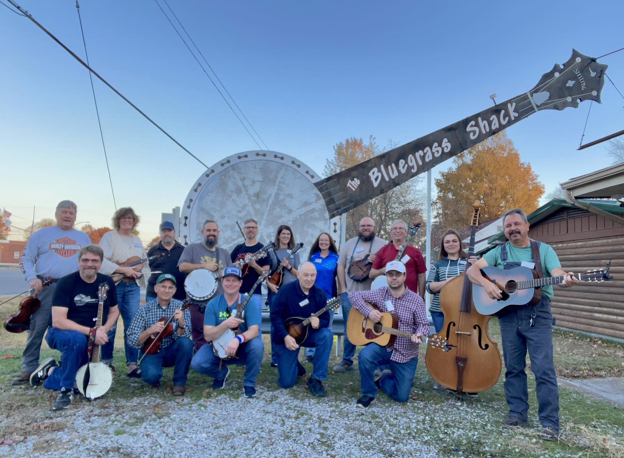 Group of people with musical instruments outdoors, a large wheel and a sign reading 'Bluegrass Shack' in the background.