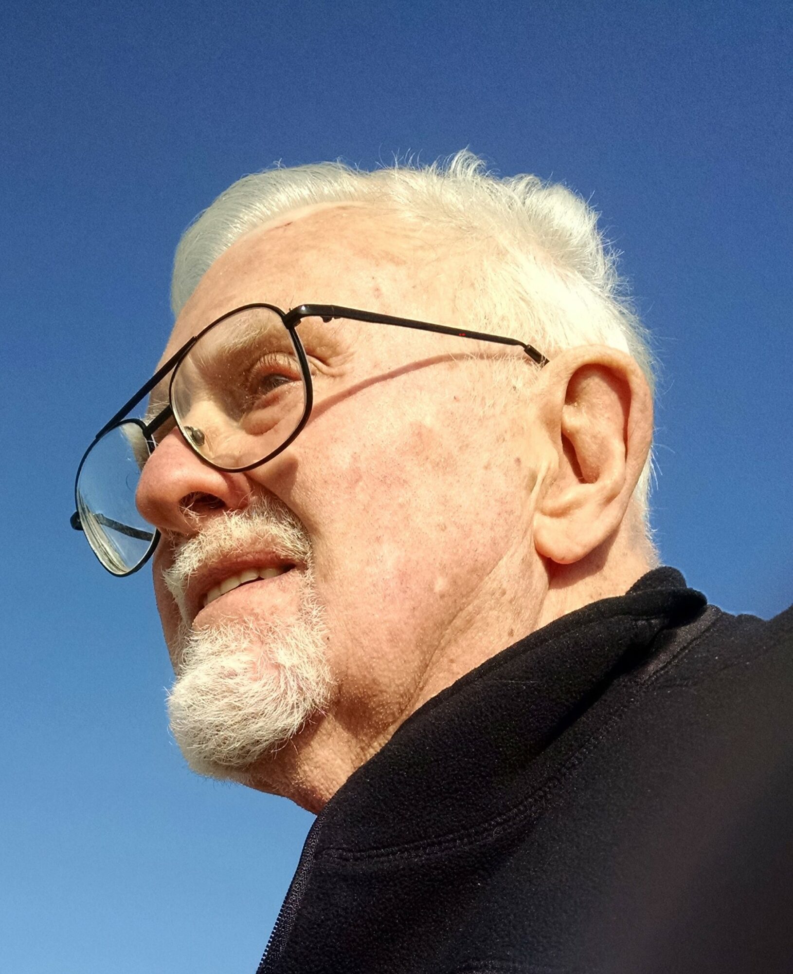Elderly man with white hair, glasses, and a beard, wearing a black jacket, outdoors against a blue sky.