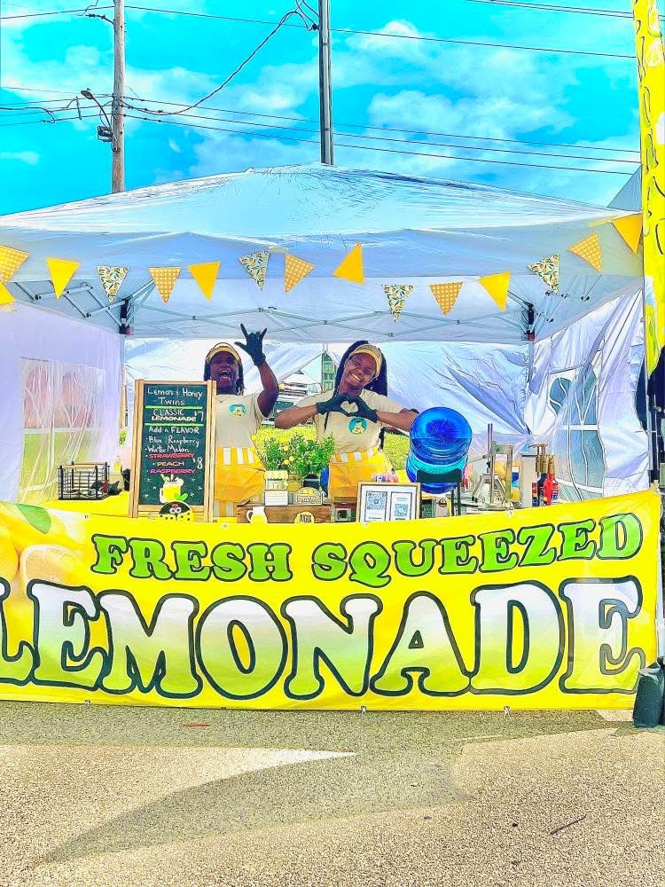 Two people behind a lemonade stand with a yellow sign reading 'Fresh Squeezed Lemonade' under a white canopy.