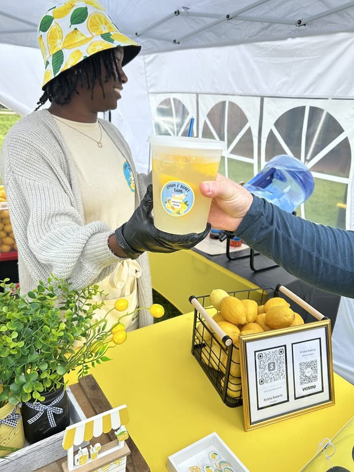 Person in yellow hat and gloves receives a cup of yellow beverage from another person at a booth.