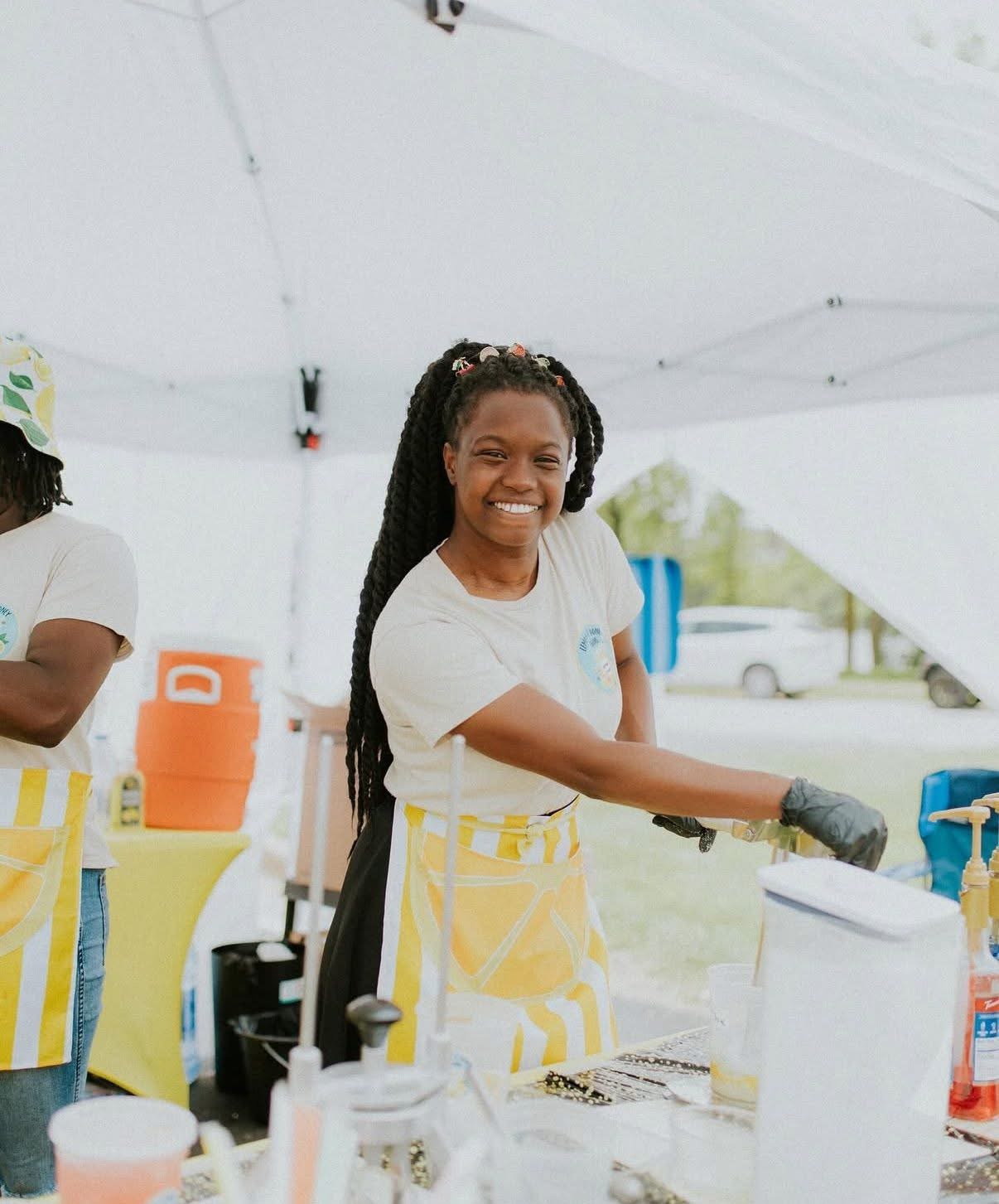 Smiling woman with long braided hair wearing a white shirt and yellow apron, serving at an outdoor event.