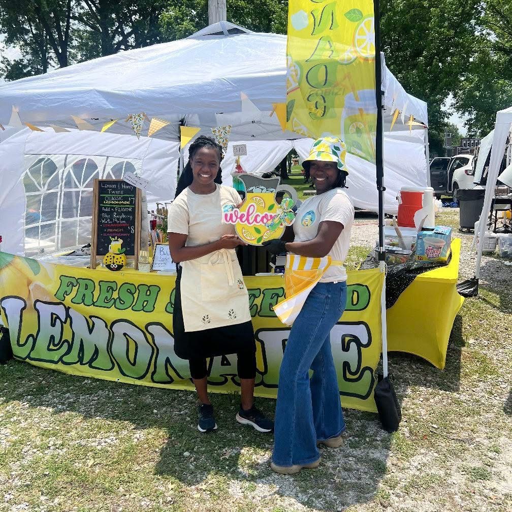 Two women standing in front of a lemonade stand, holding a pink and yellow sign that says 