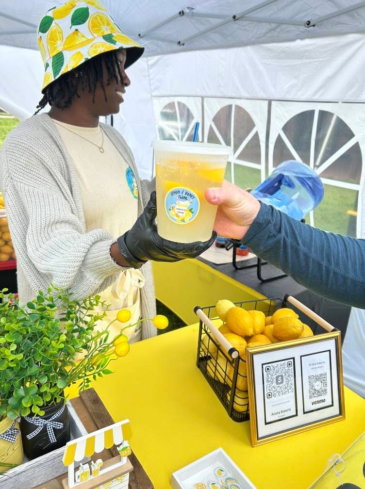 Person wearing a colorful hat receives a drink from another person at a yellow table with lemons and a QR code sign.