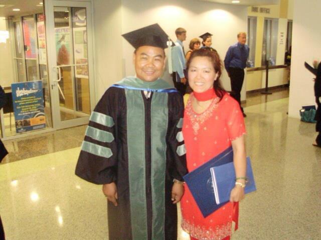 Person in graduation gown and cap standing next to woman in red dress holding a blue folder, in a lobby area.