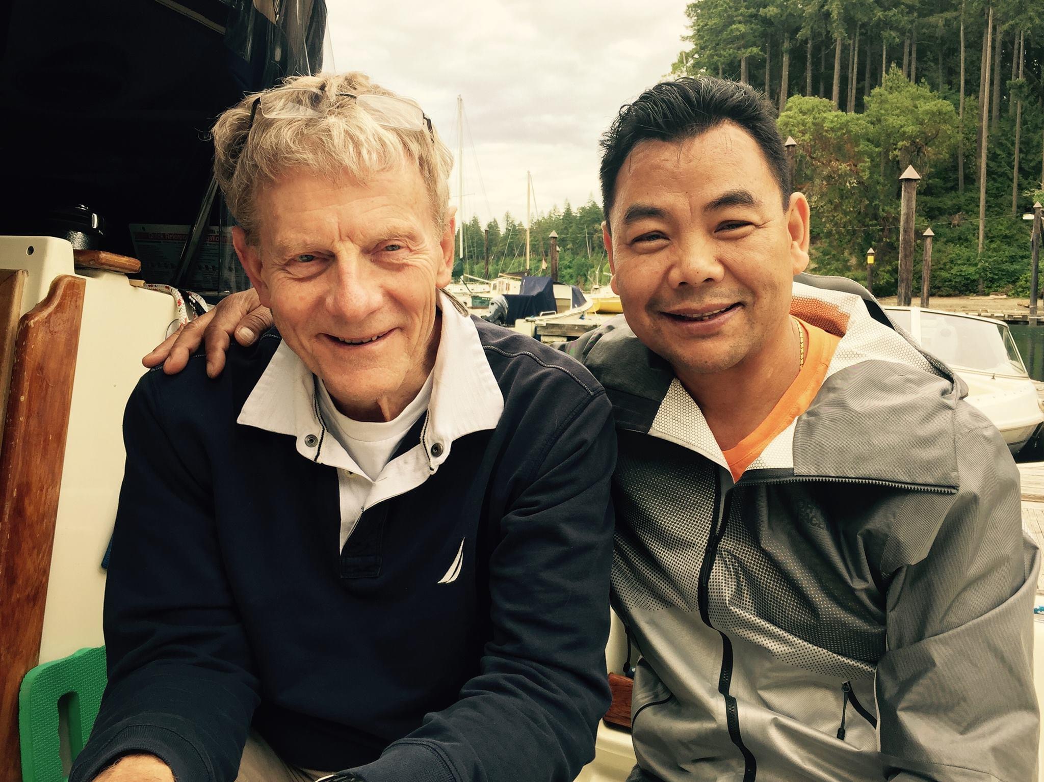 Two men sitting outdoors, smiling, with trees and boats in the background.