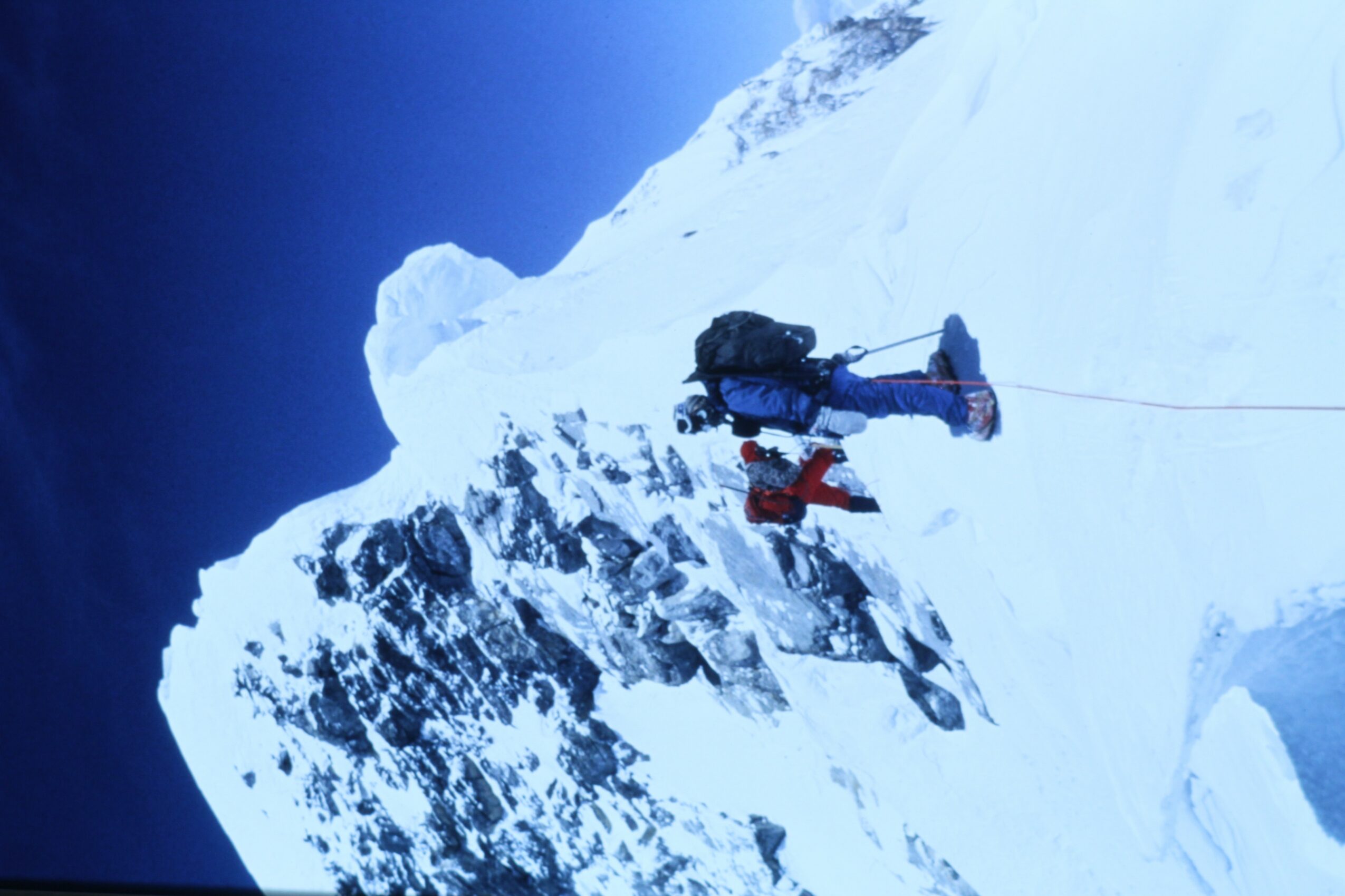 Person climbing snowy mountain with gear, surrounded by snow and ice, against a dark sky.