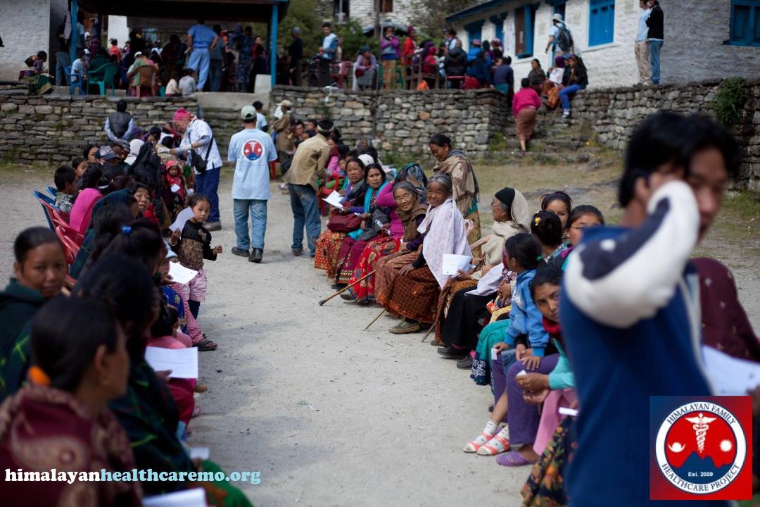 People seated and standing in a queue outside buildings, some wearing masks, in a rural setting.