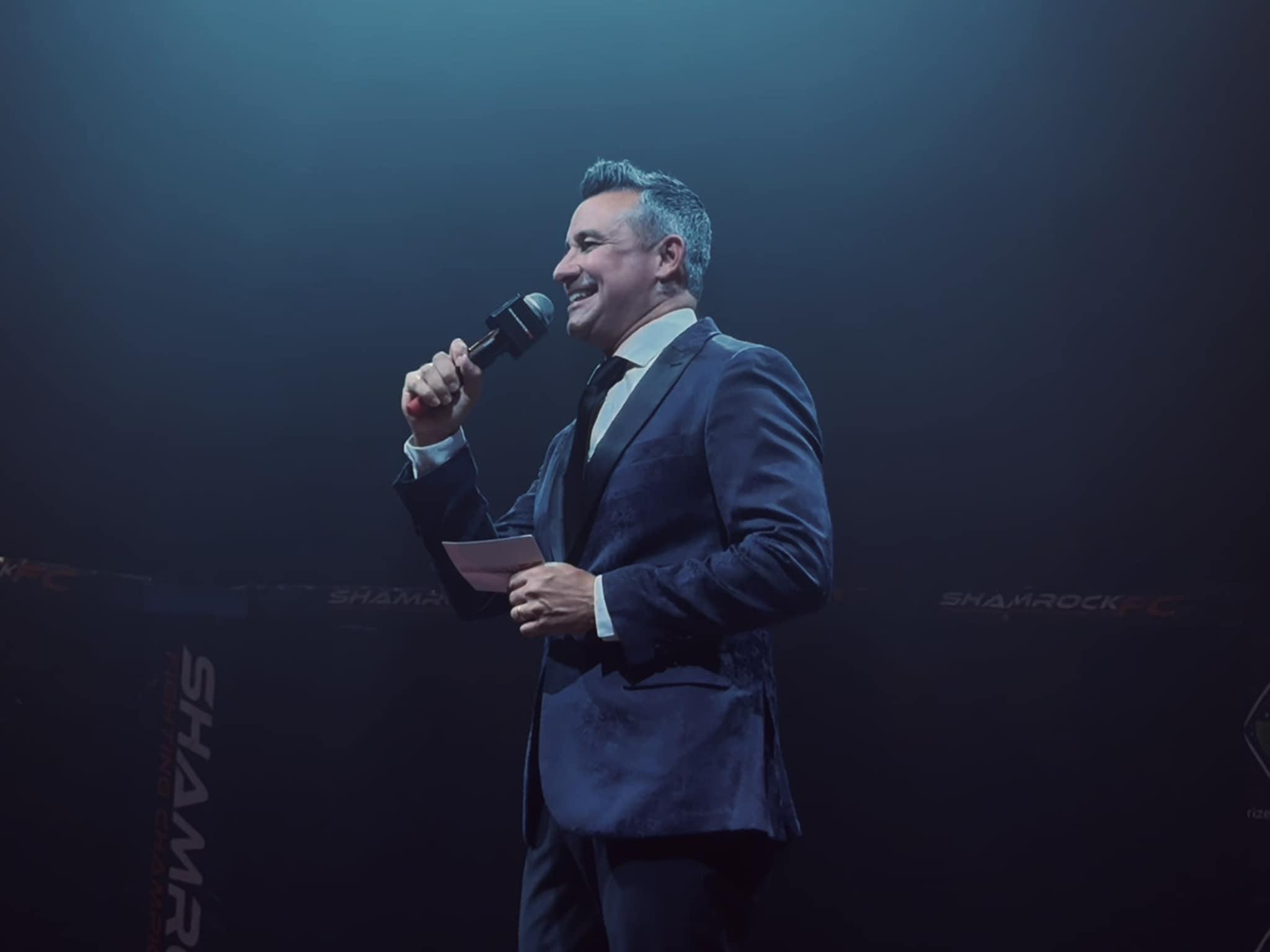 Man in a suit holding a microphone and paper, smiling on stage with dark background.