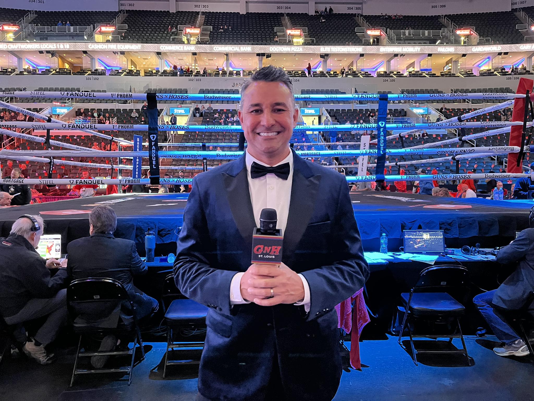 Man in tuxedo holding microphone in boxing ring with audience in background.
