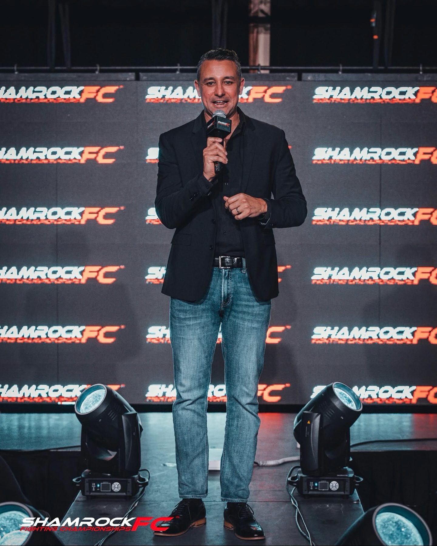 Man standing on stage with microphone, smiling, in front of Shamrock FC backdrop, wearing a dark blazer and jeans.